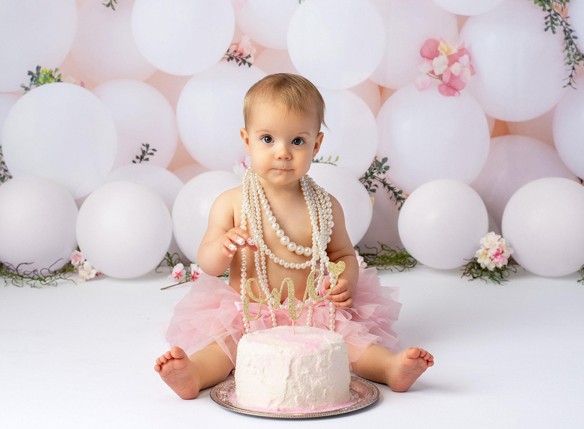 baby girl wearing layered pearl necklace and pink tutu sitting in front of cake with gold glitter ONE sign with white balloons in background and frosting on fingers