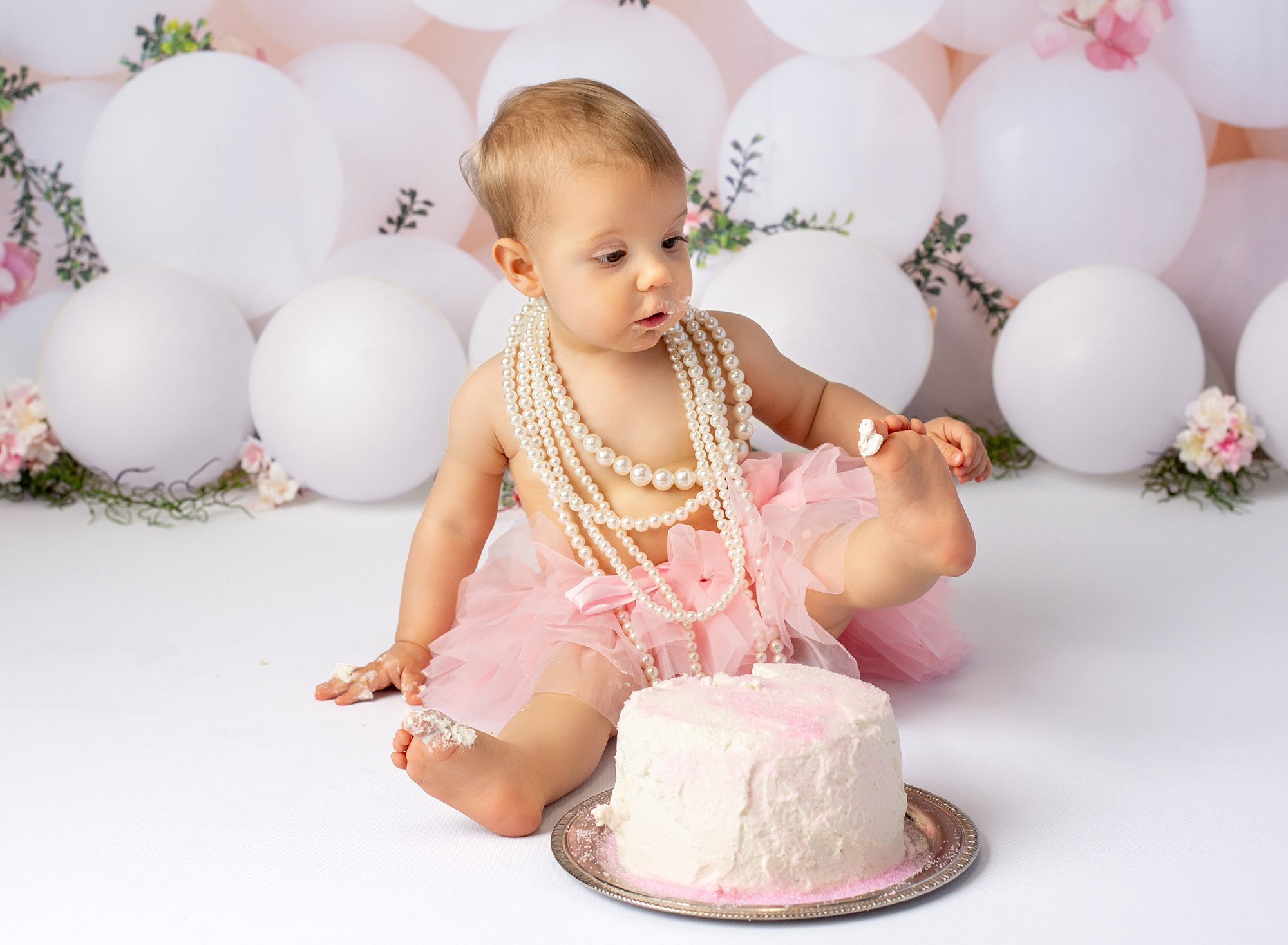 1 year baby photoshoot baby girl wearing layered pearl necklace and pink tutu sitting in front of cake holding up foot with frosting on toe with white balloons in background