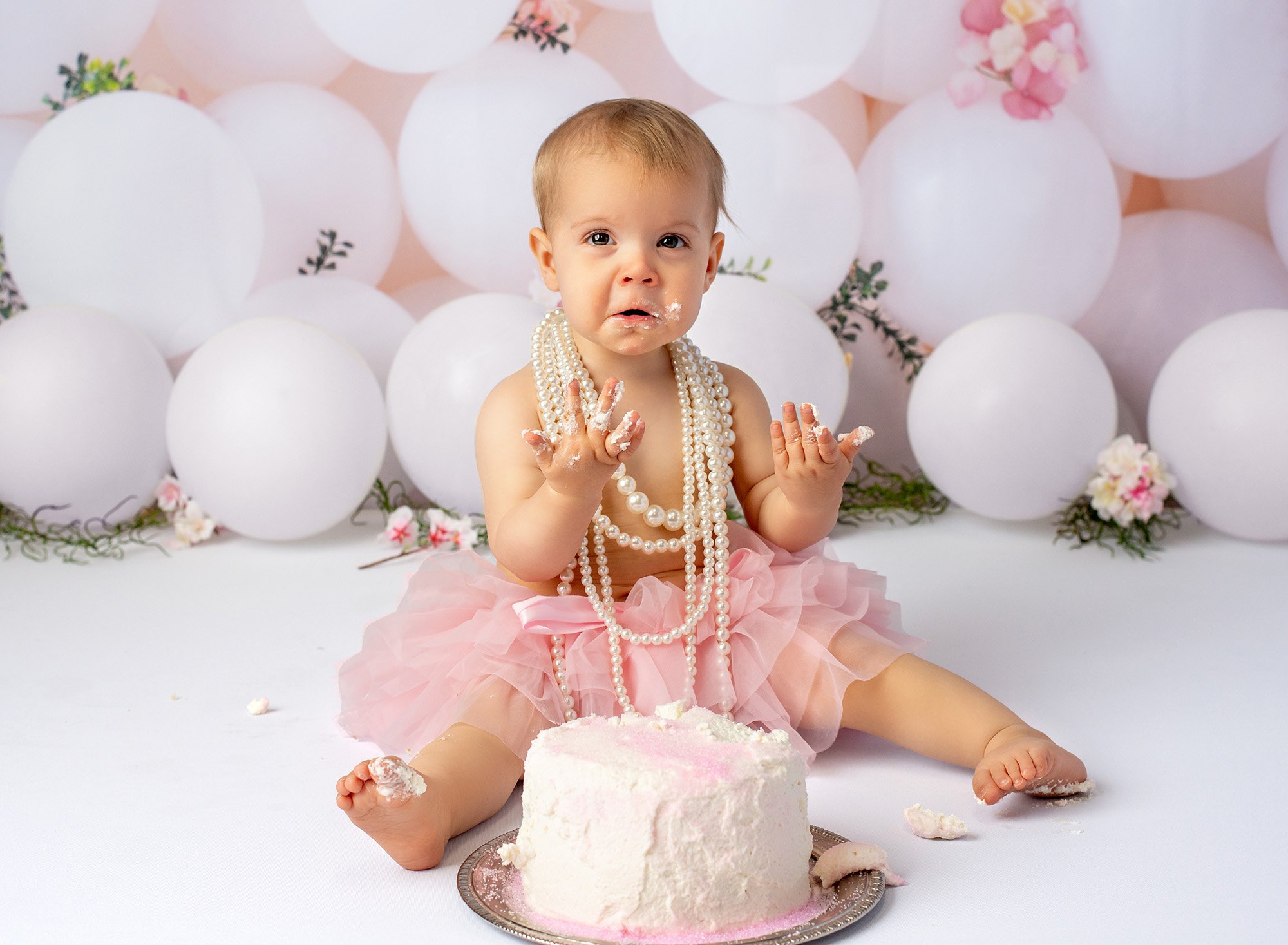 baby girl wearing layered pearl necklace sitting in front of cake with frosting on hands with white balloons in background