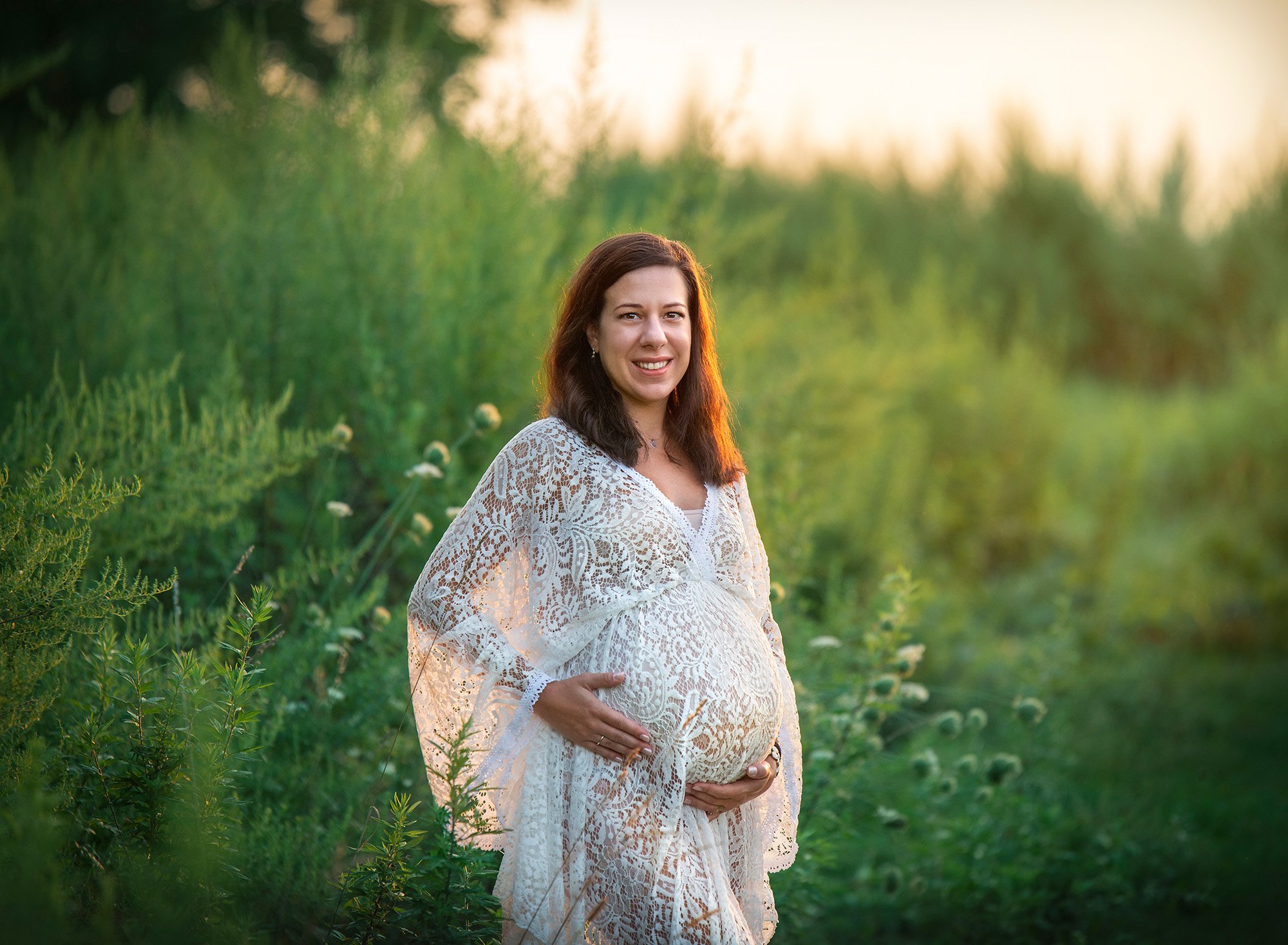 pregnant woman wearing bohemian laced dress holding her stomach while standing in a field at sunset