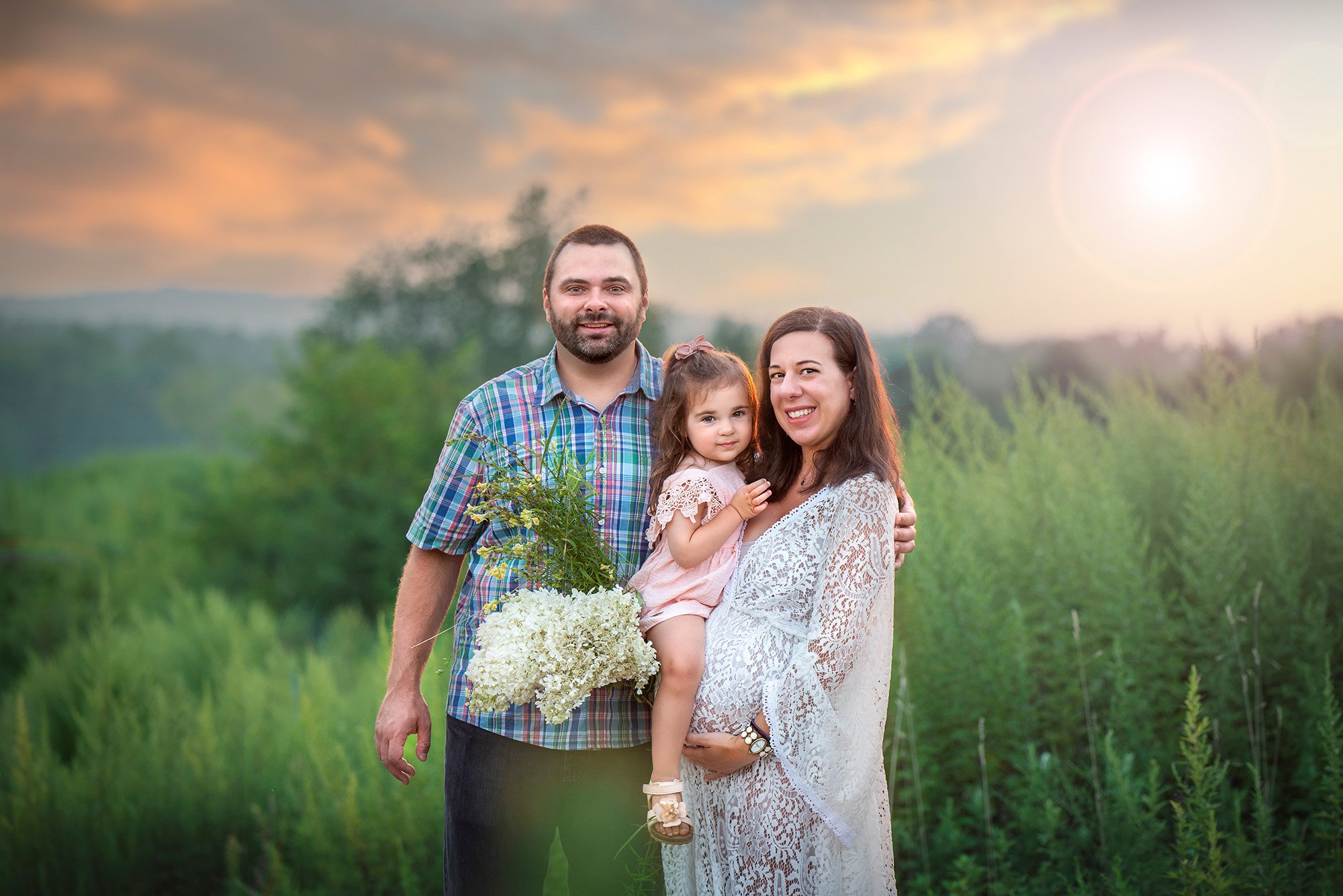 pregnant woman and partner posing in a field at sunset holding their daughter