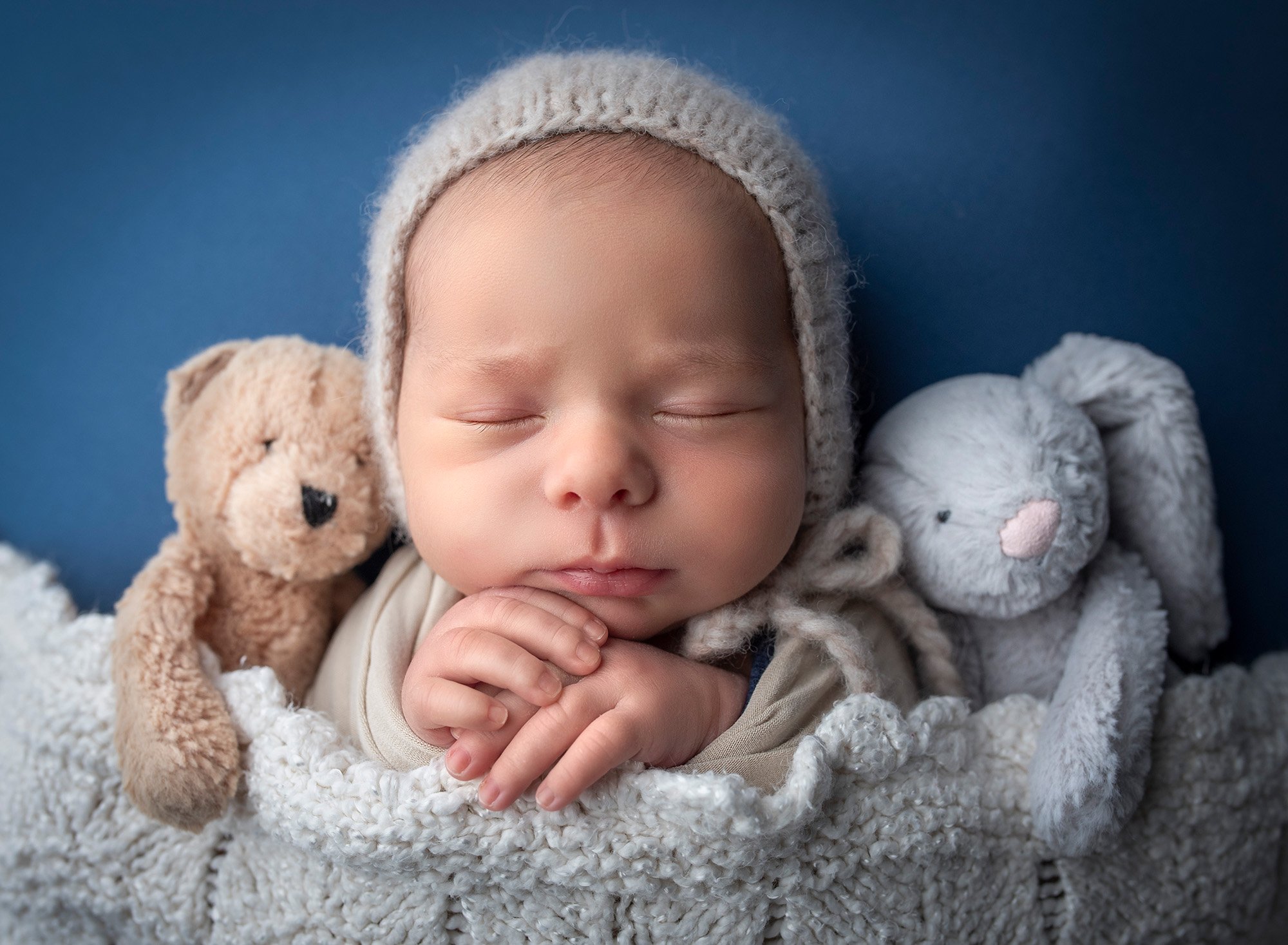 newborn baby boy wearing a bonnet snuggled in between a teddy bear and a bunny