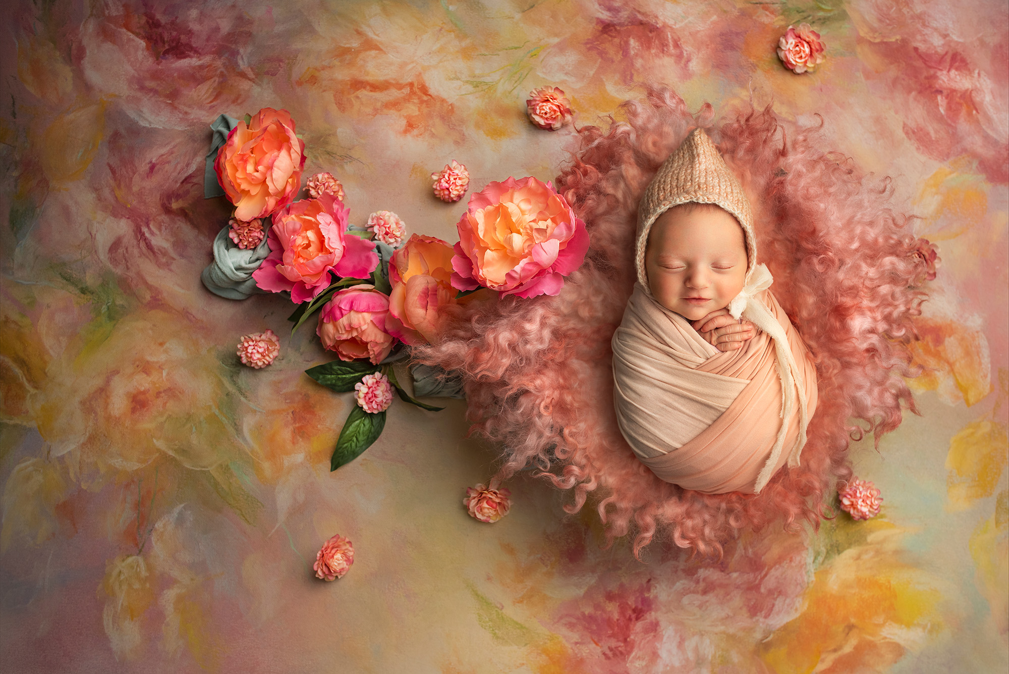 Adorable smiling newborn wearing a bonnet, wrapped in warmth on a vintage rose backdrop of oranges, pinks, and yellows.