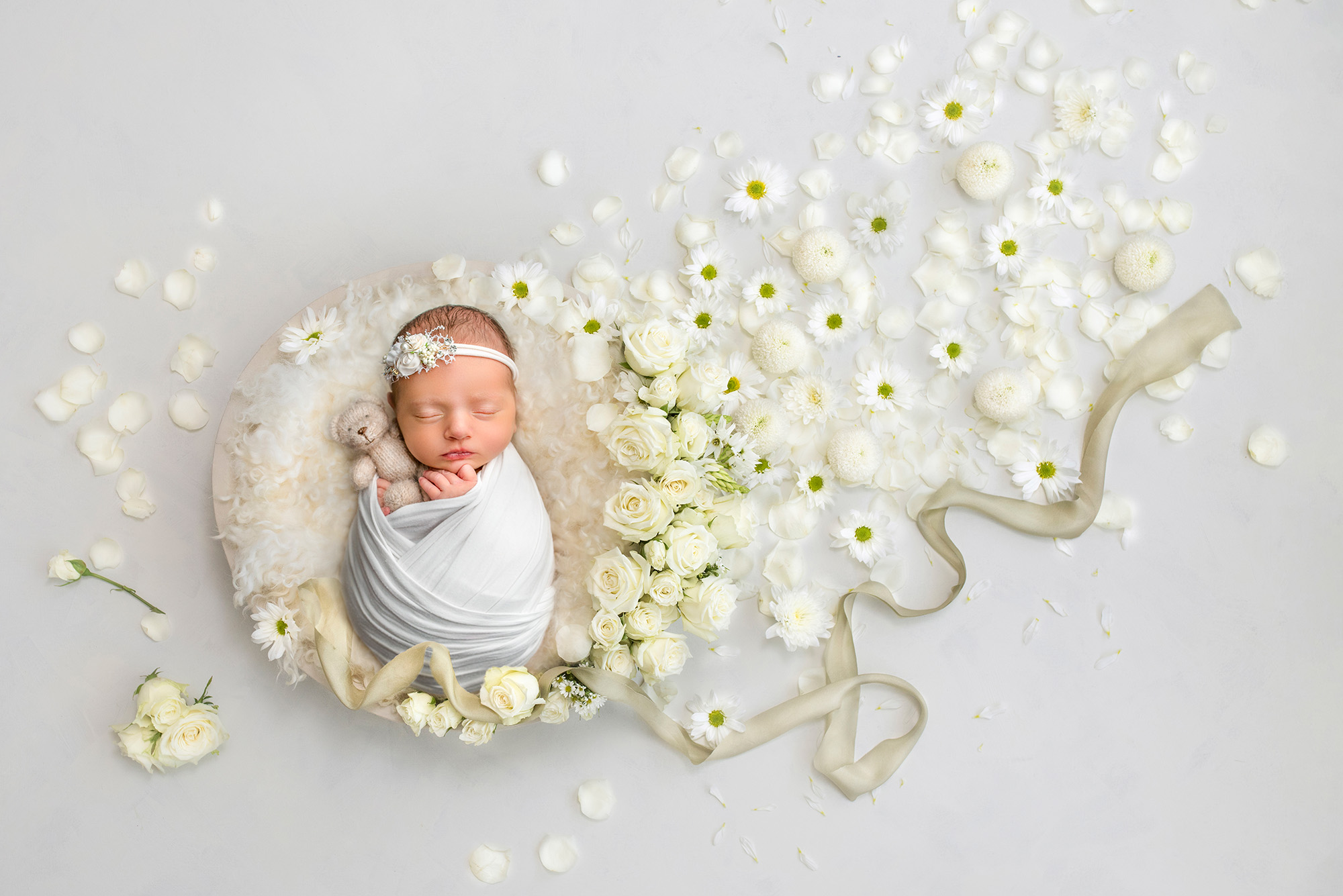 Wrapped in white and cradling a small teddy bear, this baby lies in a cream bowl surrounded by daisies and khaki ribbon, a picture of tranquility