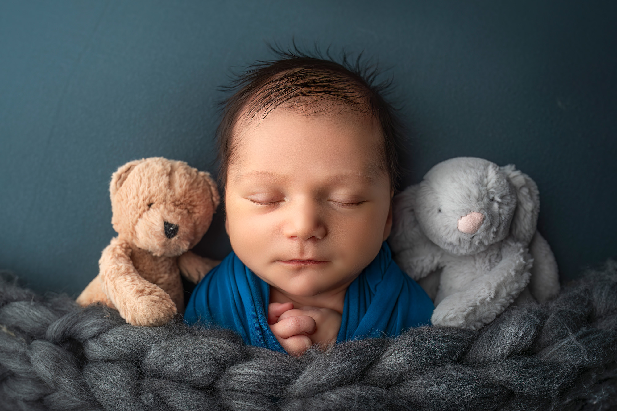 Newborn boy sleeping in between a stuffed teddy bear and bunny, under a grey blanket