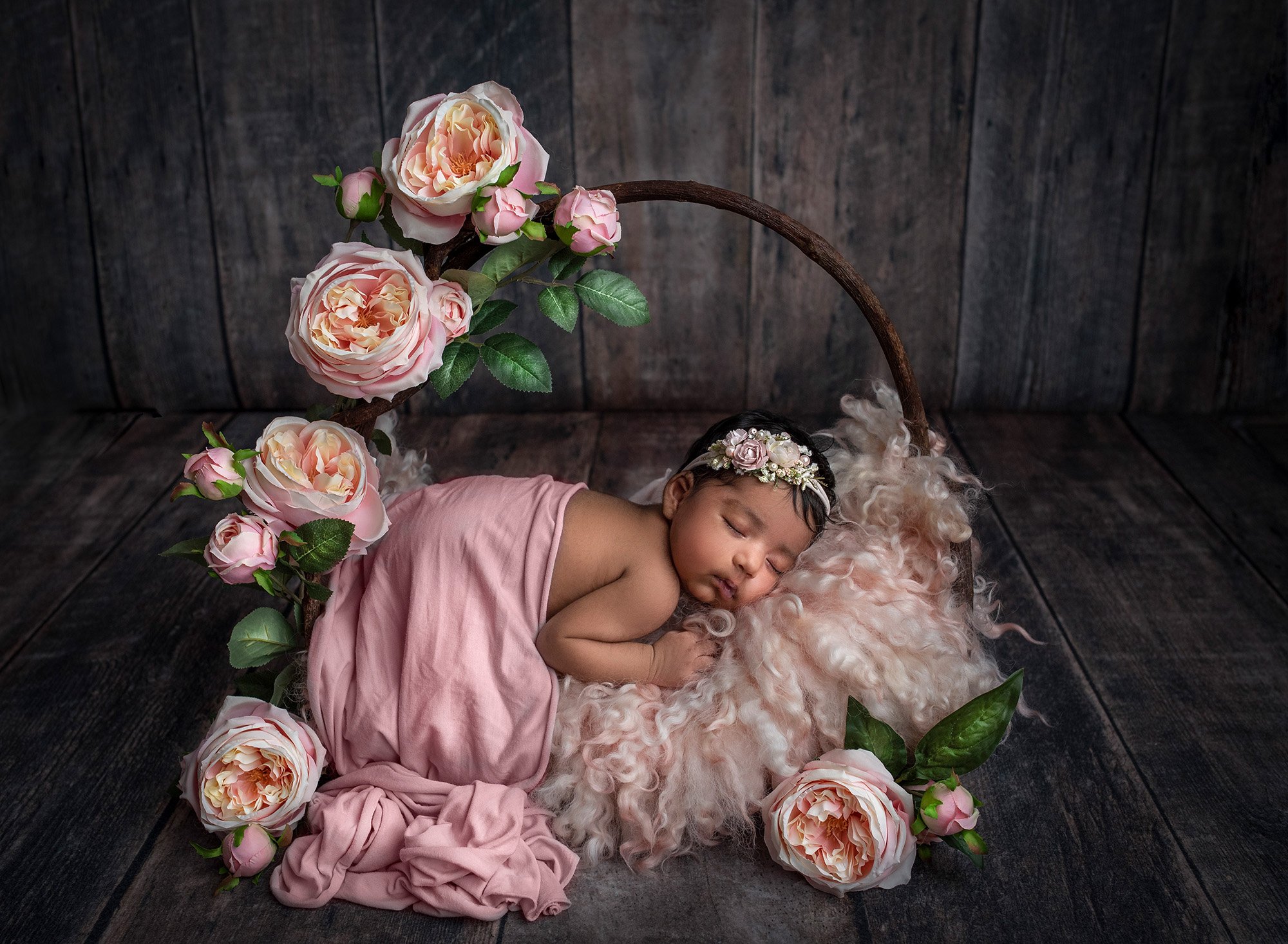Newborn Girl Photography newborn baby girl draped in pink sleeping on top of rustic wooden bed with pink fuzzy blanket surrounded by pink flowers on wooden background