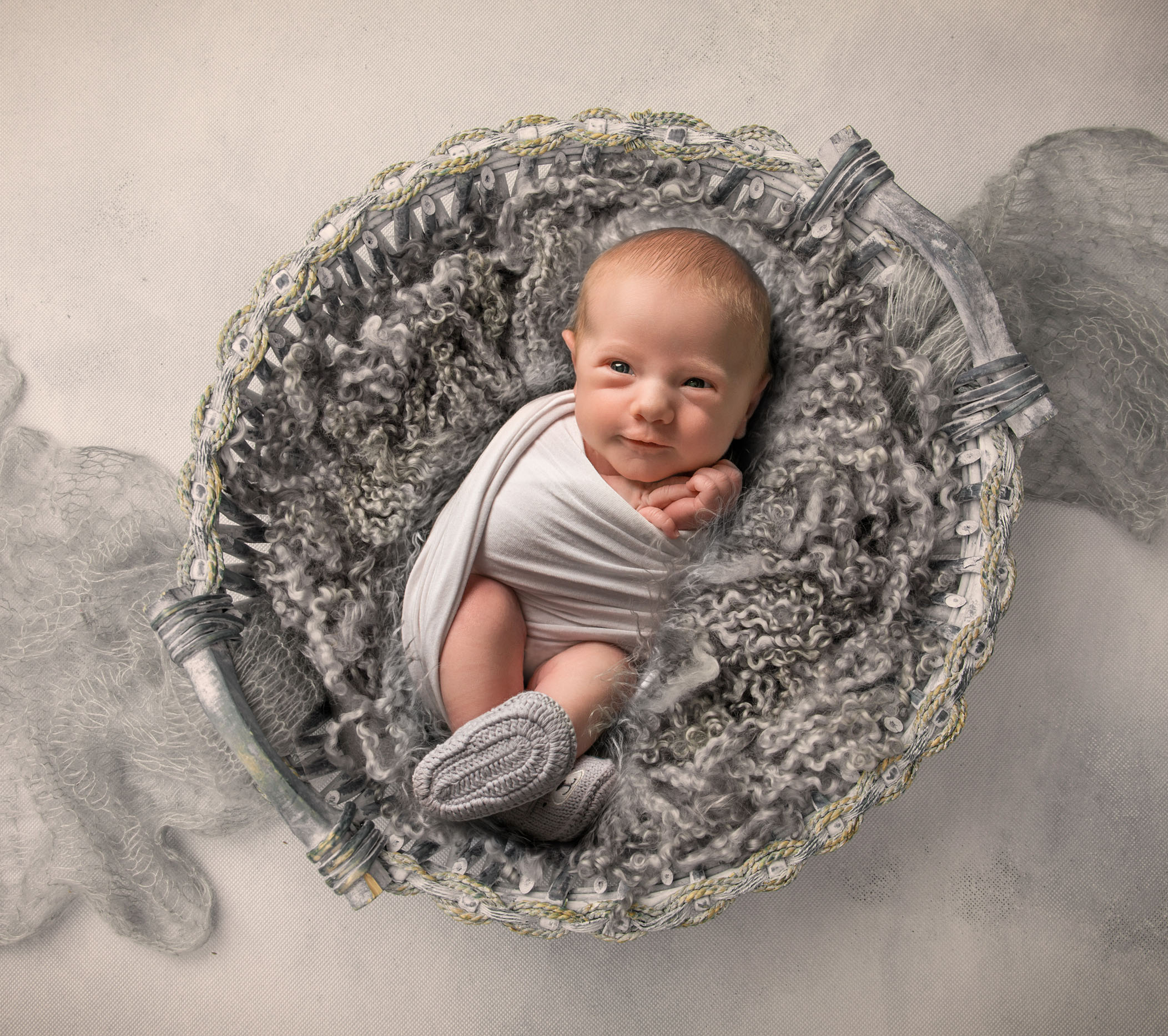 newborn baby boy swaddled in grey fluff basket with grey booties on his feet