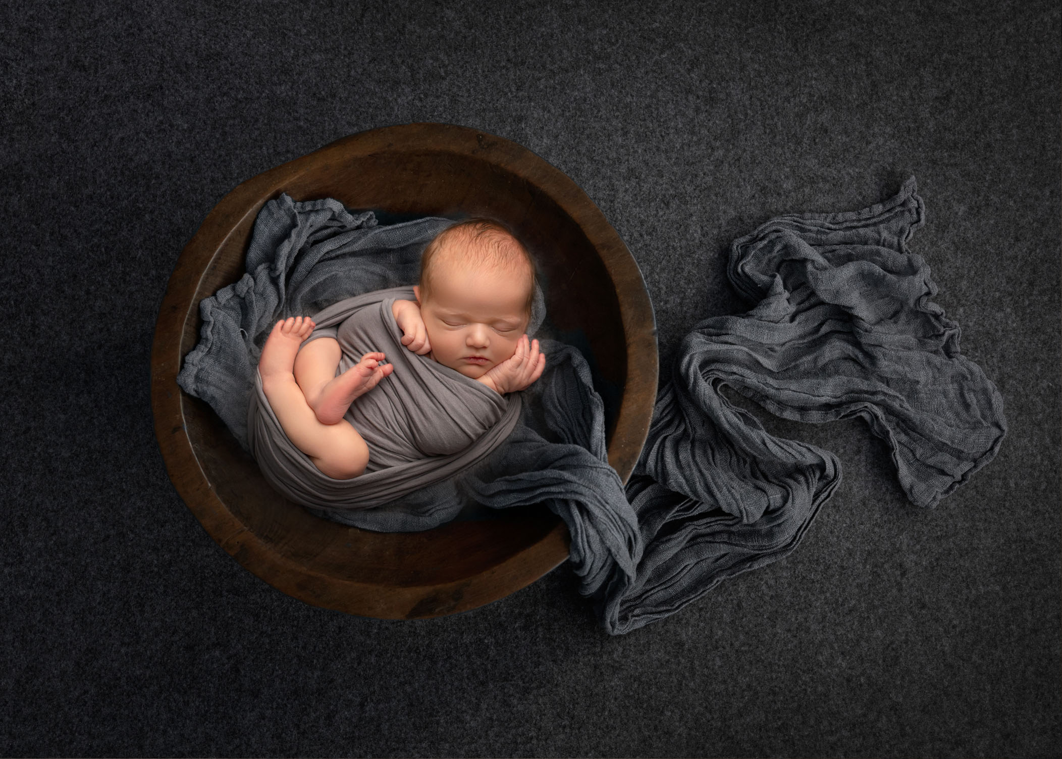 Swaddled newborn boy sleeping in wooden bowl lined with grey wrappings