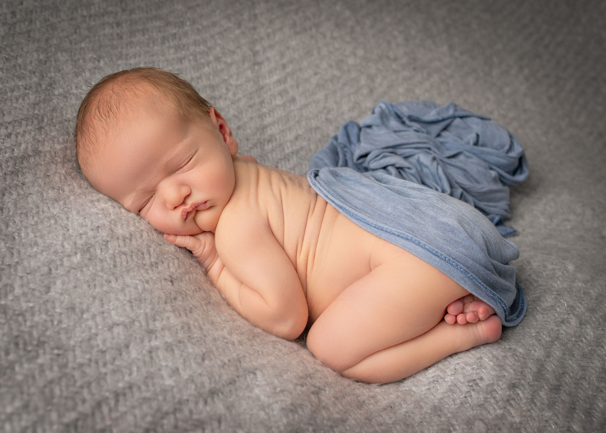 Newborn baby boy sleeping on grey blanket in bum up pose with blue wrap over his bottom