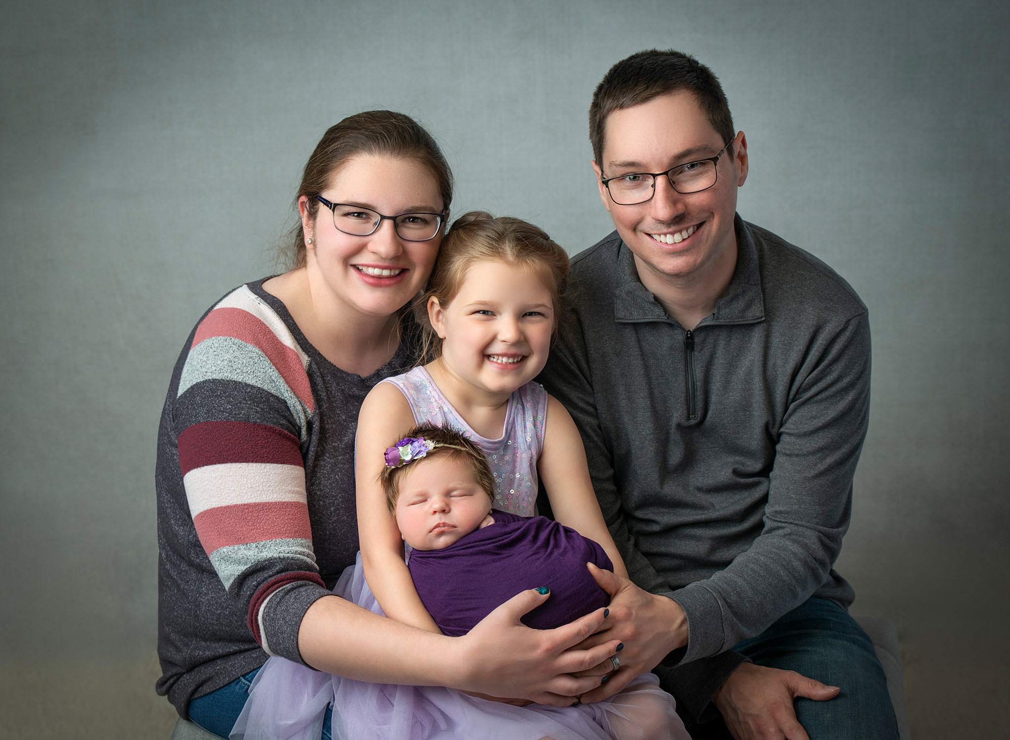 couple posing with younger daughter and newborn baby girl swaddled in purple