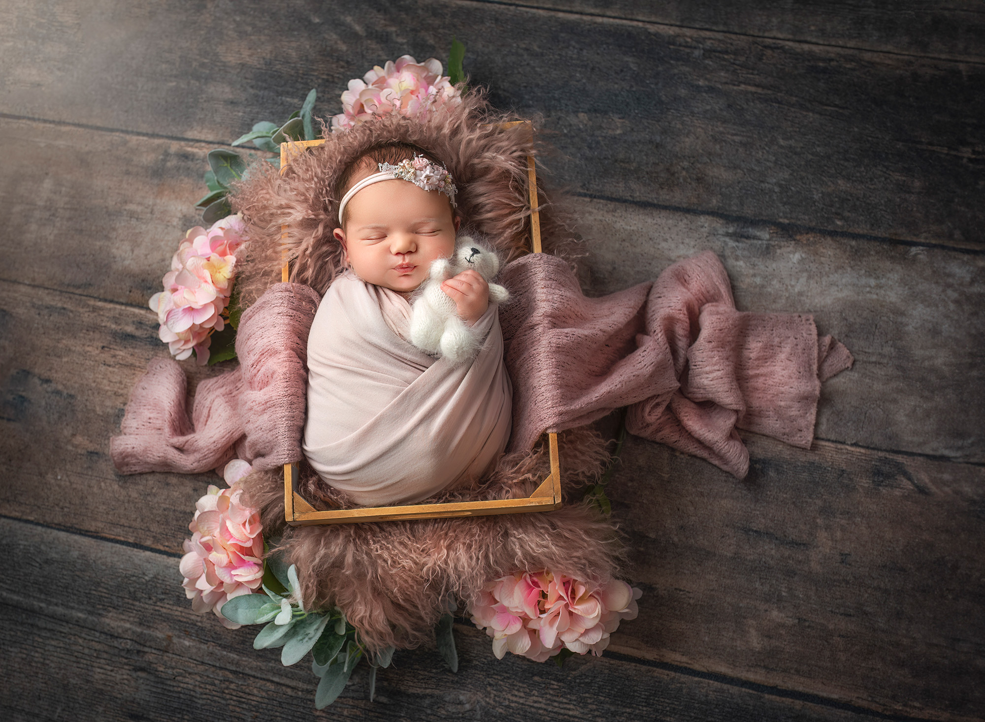 Newborn girl resting in a crate, cuddling a fuzzy white teddy bear amidst soft pink furs