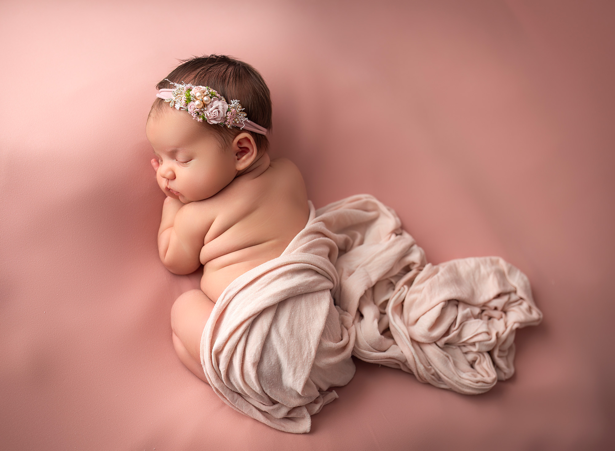 Newborn girl wearing a dainty pink pearled headband as she sleeps peacefully on a pink backdrop