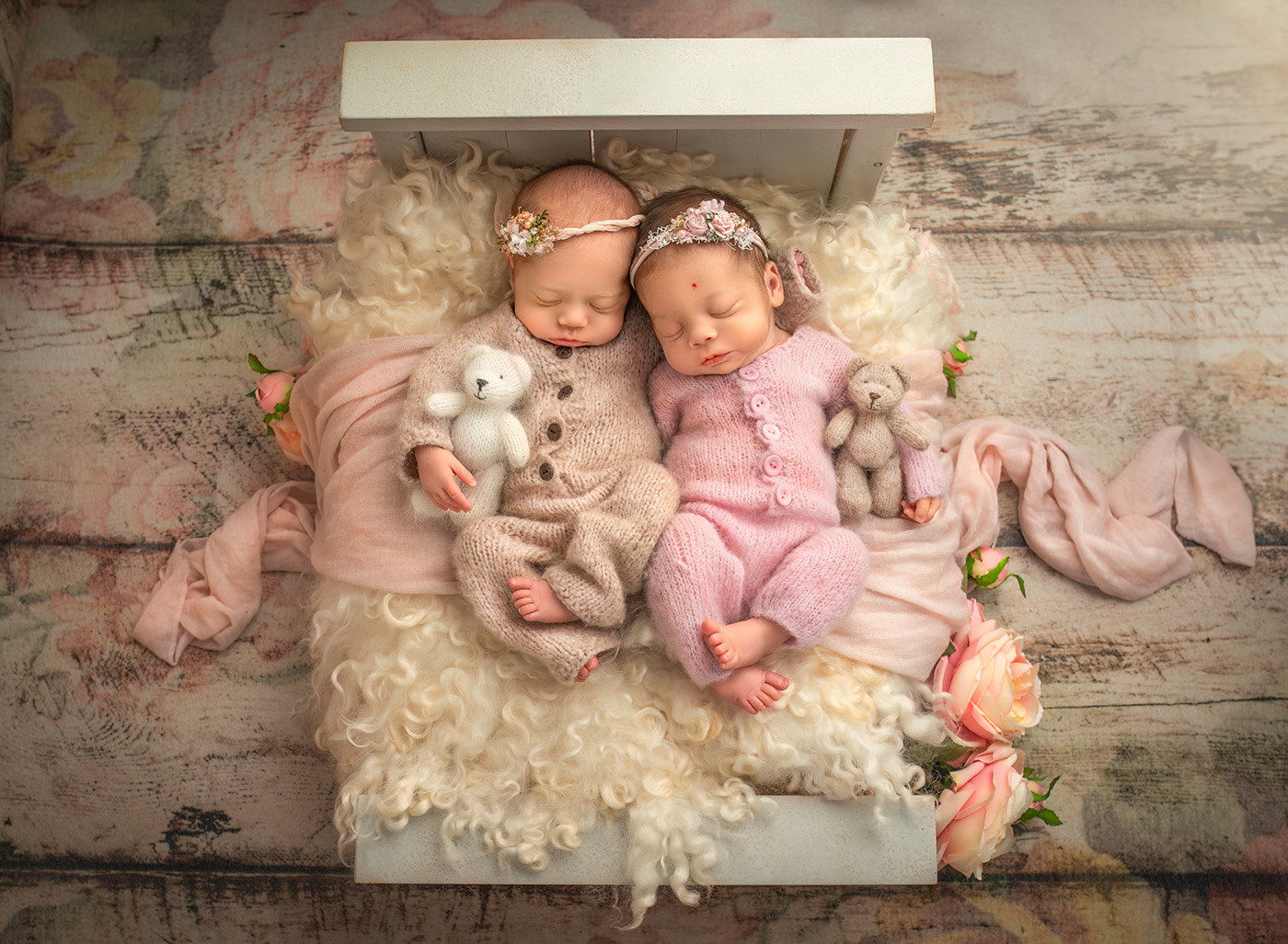 newborn twin photography two twin baby girls snuggled in knitted rompers with knitted teddy bears sleeping on a tiny bed with cream wool fluff and pink roses