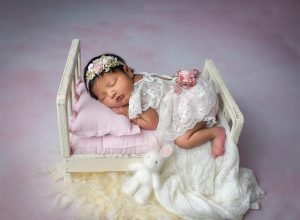 newborn baby girl wearing laced dress asleep on a rustic miniature bed
