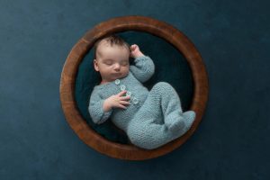 6 week old newborn boy wearing knitted romper asleep in a wooden bowl on teal background