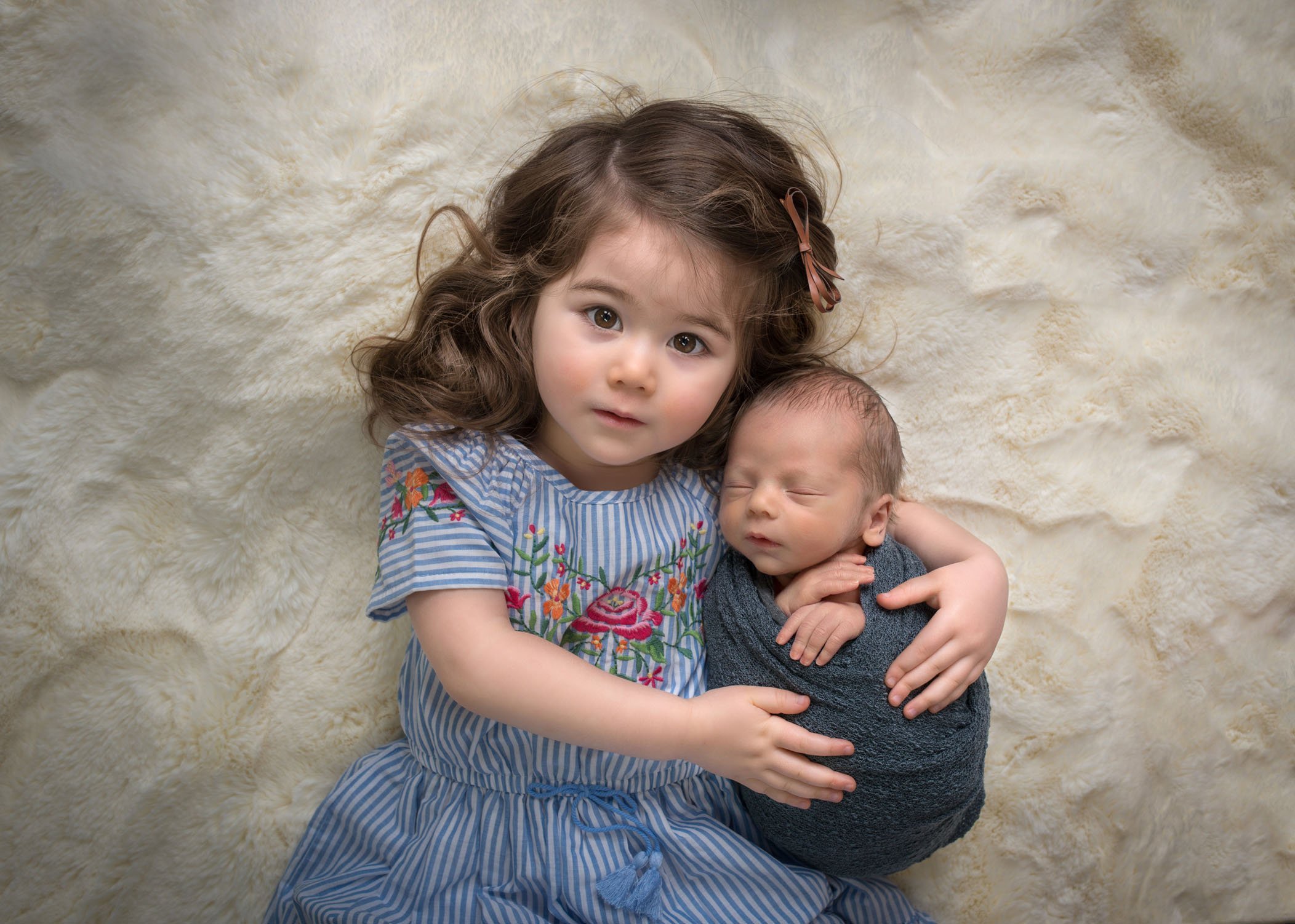 2 year old curly haired sister holding her newborn brother on cream fur