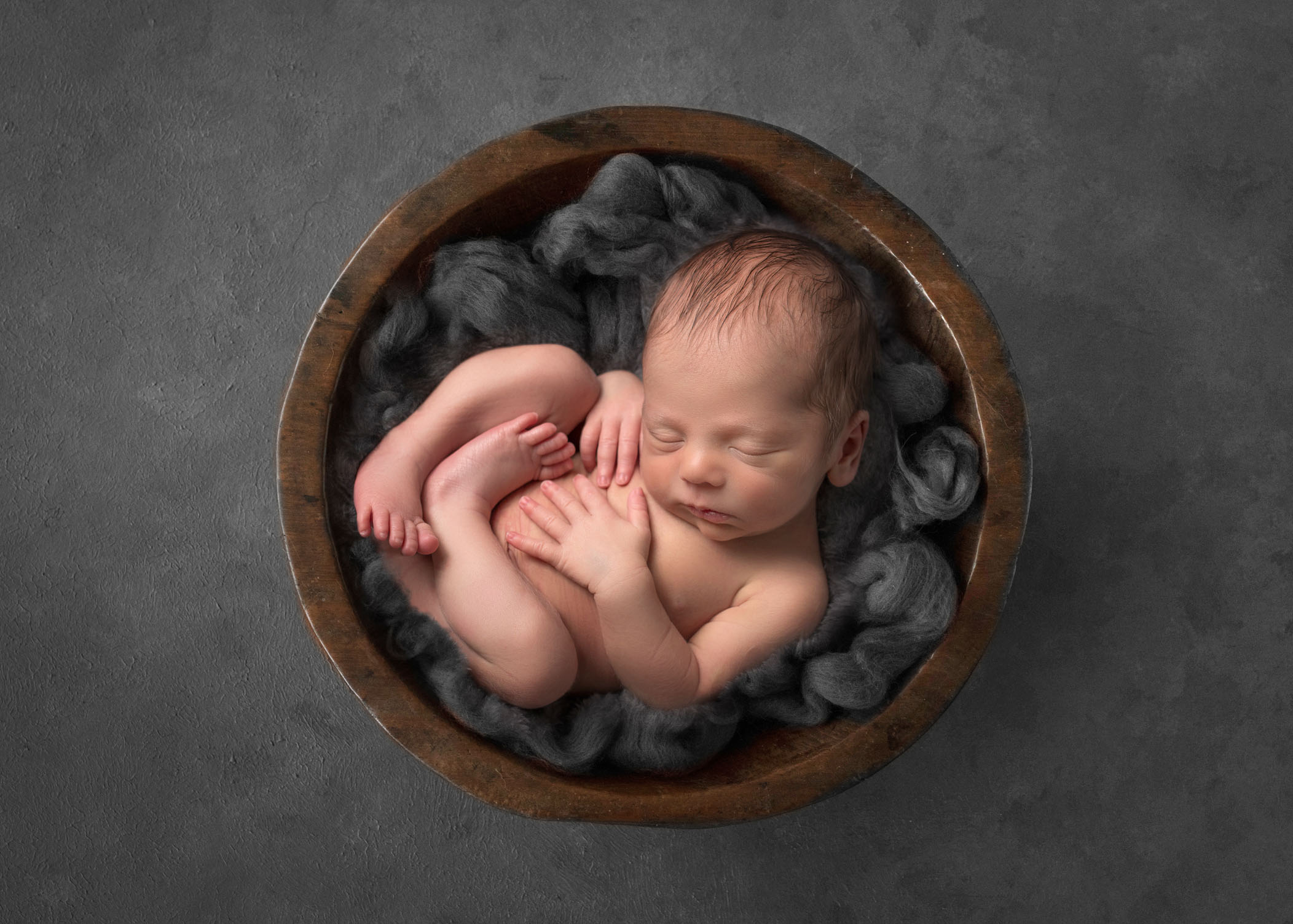 newborn baby boy curled up in a wooden bowl of grey fluff