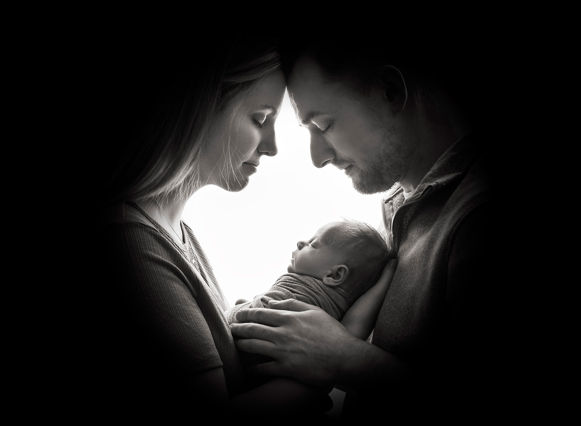 couple with eyes closed holding newborn baby in a black and white silhouette