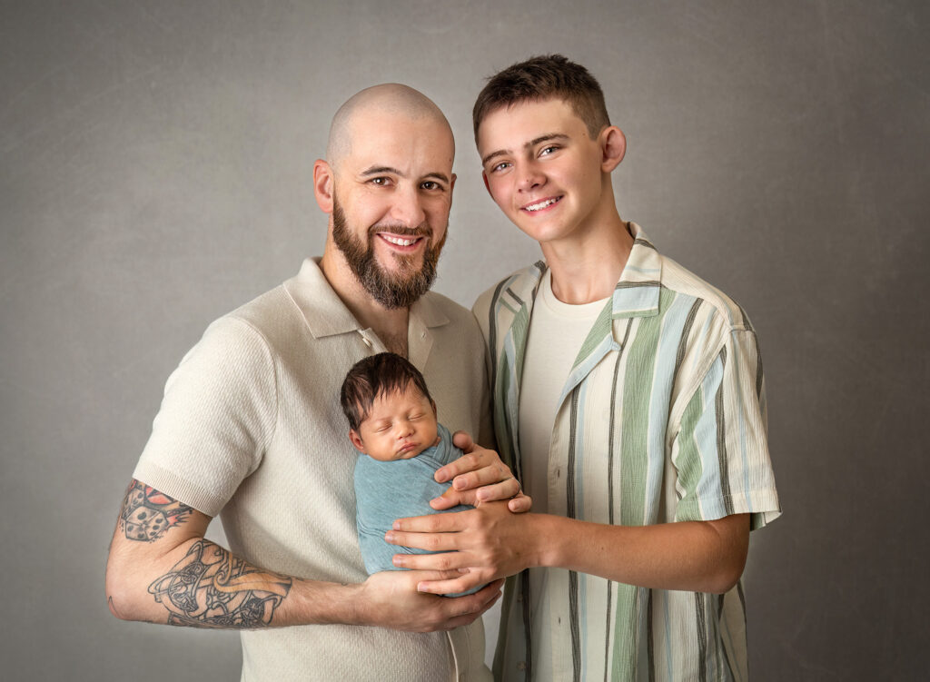 Dad and big brother smiling while safely holding a swaddled newborn in sage during a studio portrait.