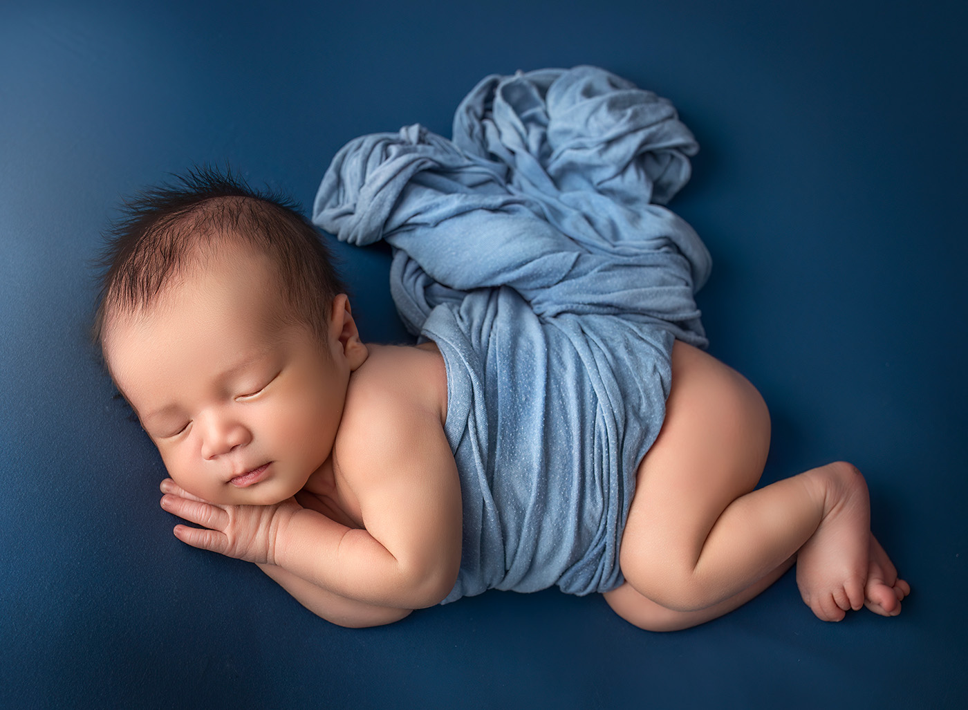 vibrant newborn photos baby boy sleeping on a deep blue background with a light blue wrap over his tummy on his side with his hands under his cheek