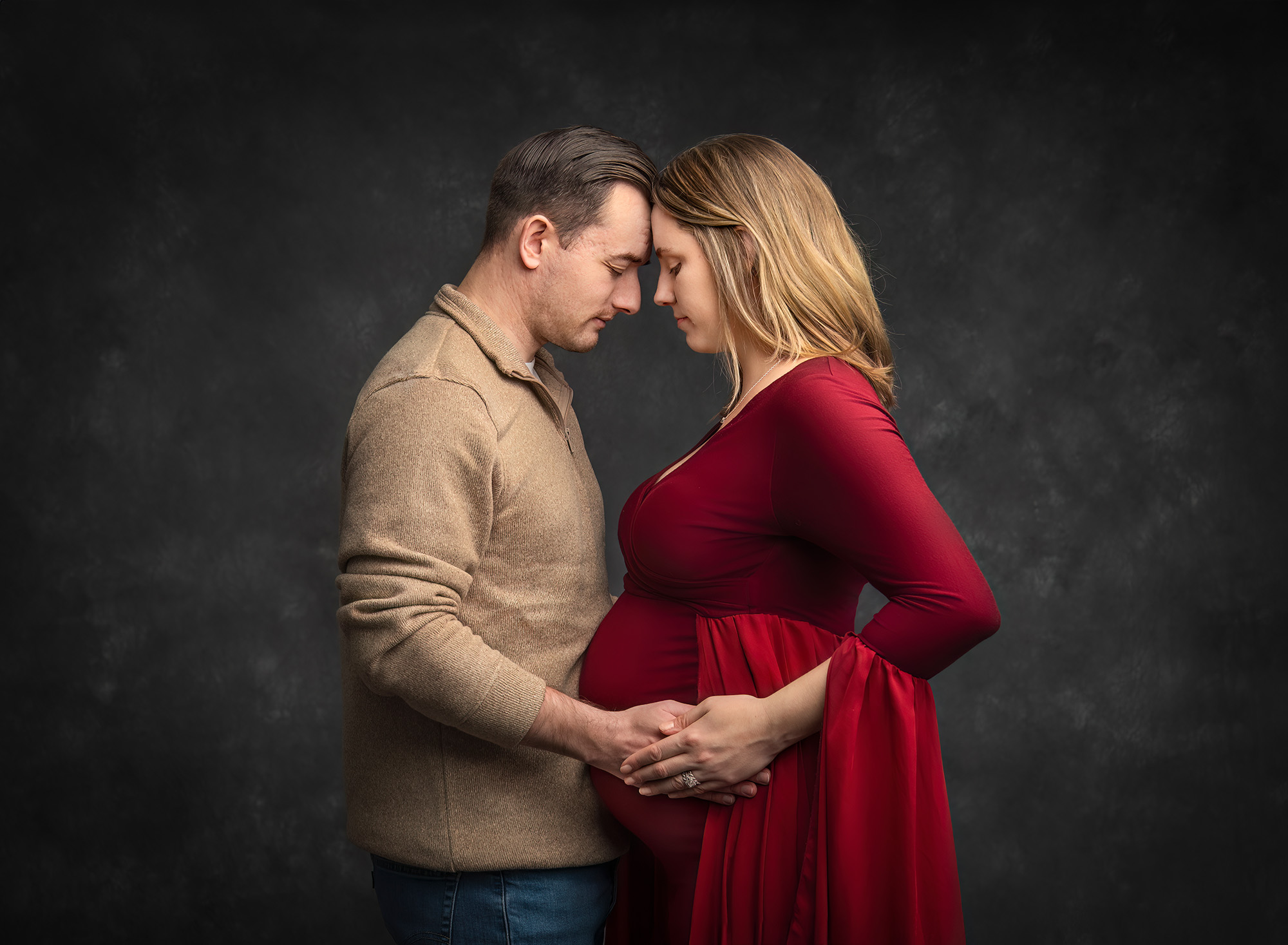 blonde woman holding hands with husband cradling pregnant belly in a cranberry dress