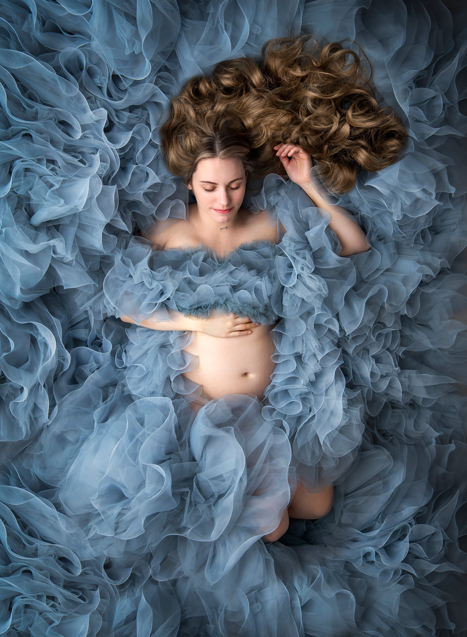 blonde curly haired woman laying down exposing pregnant belly in a large blue ruffle gown