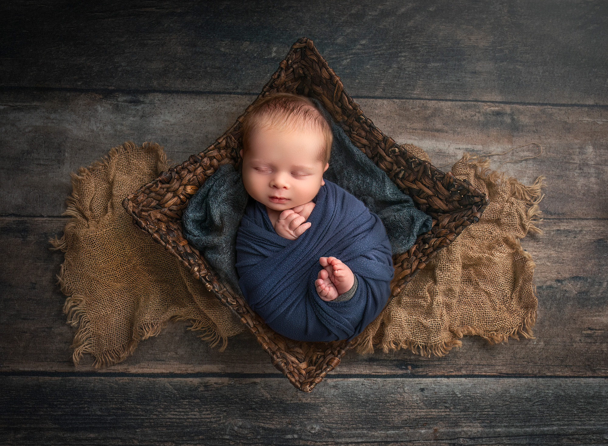 Newborn baby boy fast asleep in a wicker basket on a wooden floor, with navy blue setting the tone