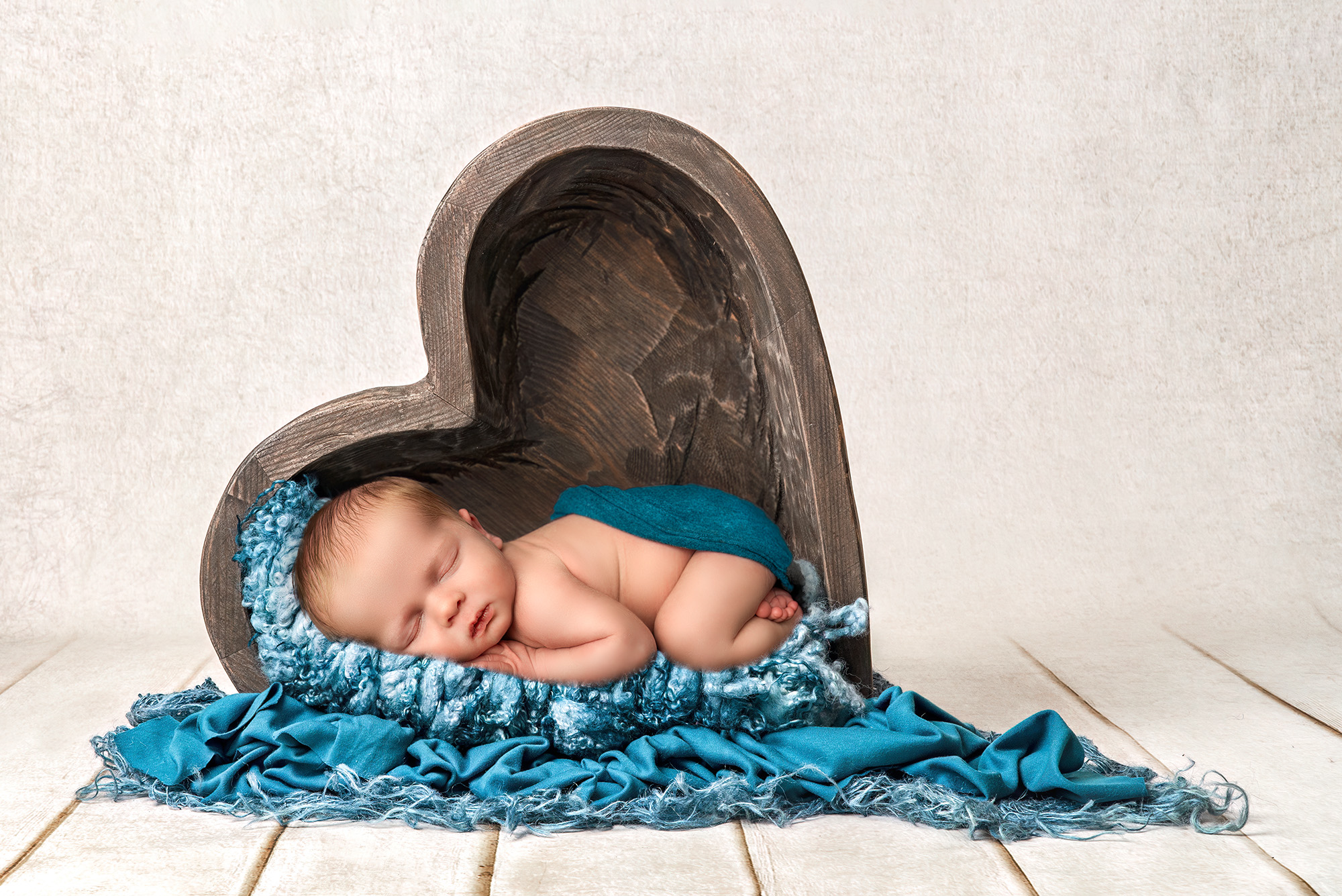 Newborn baby boy asleep in a wooden heart-shaped bowl, with textured blue blankets