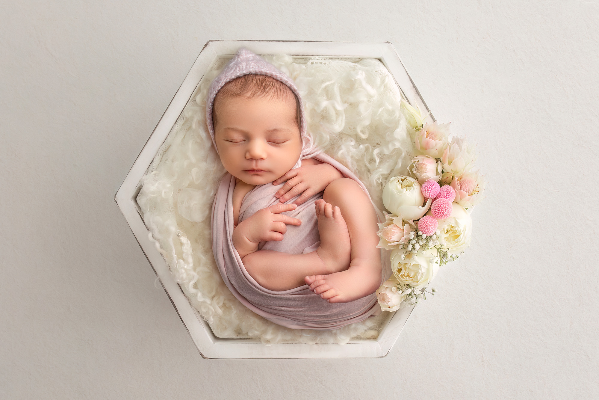Baby girl laying in a white wooden bowl in as pastel colored wrap, next to pale colored florals.