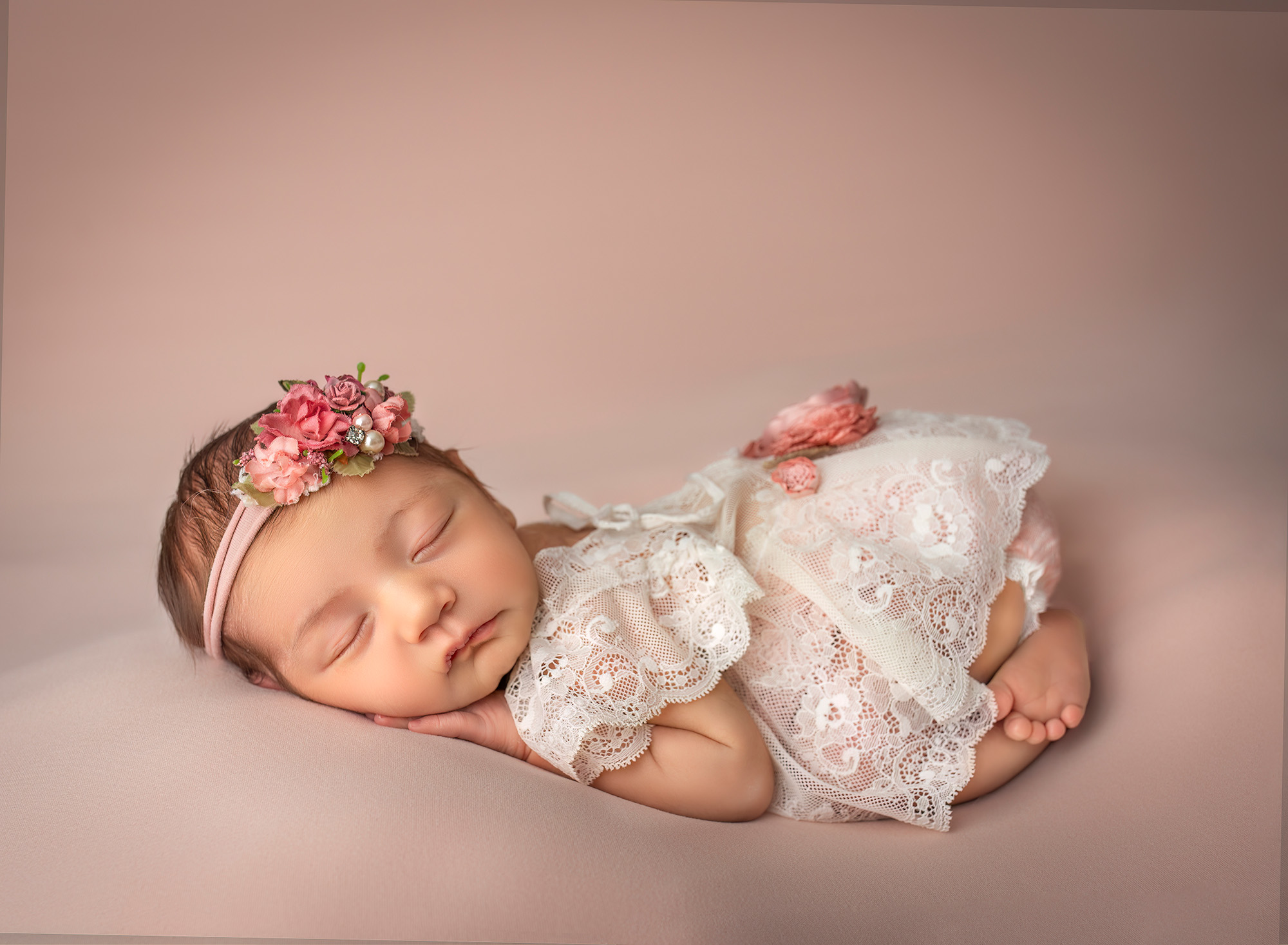 Newborn baby girl wearing a laced dress and floral headband, sleeping on her tummy