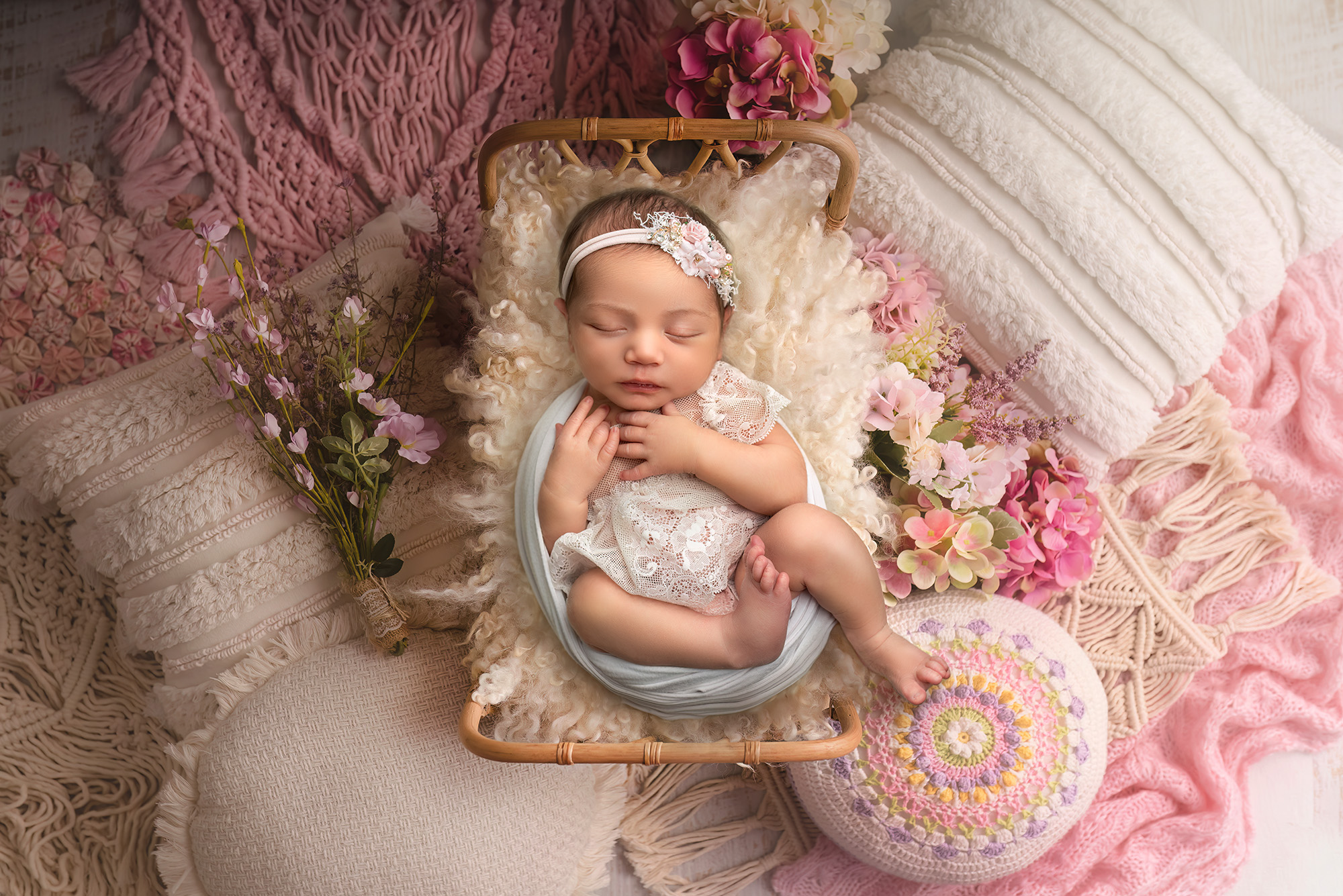 Newborn baby girl sleeping in a rattan bed, wrapped in lace, surrounded by pillows and crochet