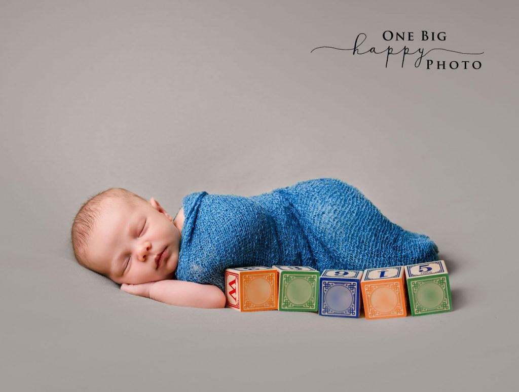 Newborn baby lying on grey background wrapped in blue blanket with name blocks in front of him
