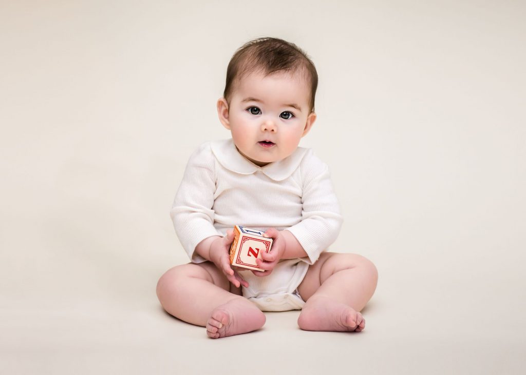 6 month old baby sitting up holding a block on a cream background