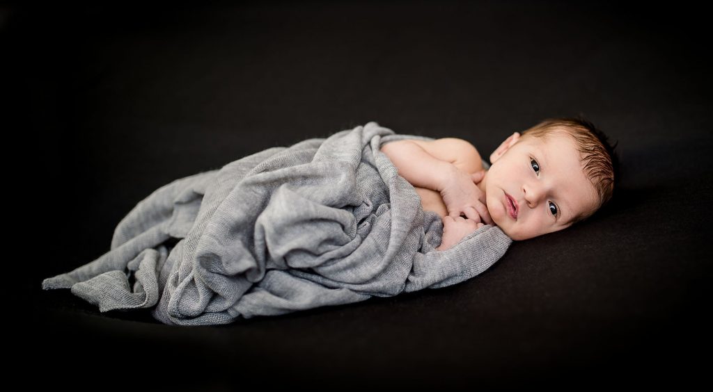 awake newborn lying on dark grey background wrapped in light grey scarf
