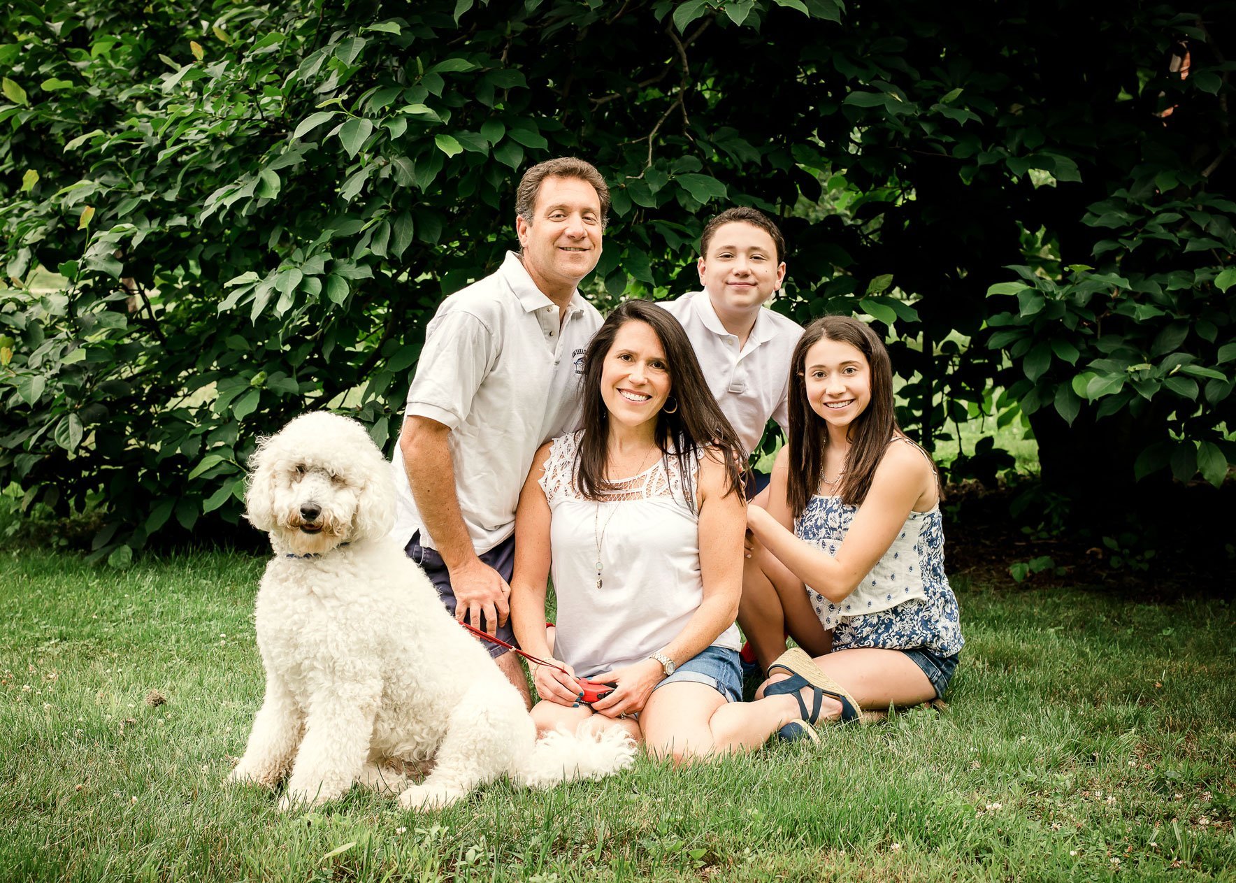 family of 5 pose outside in summer with their giant white poodle