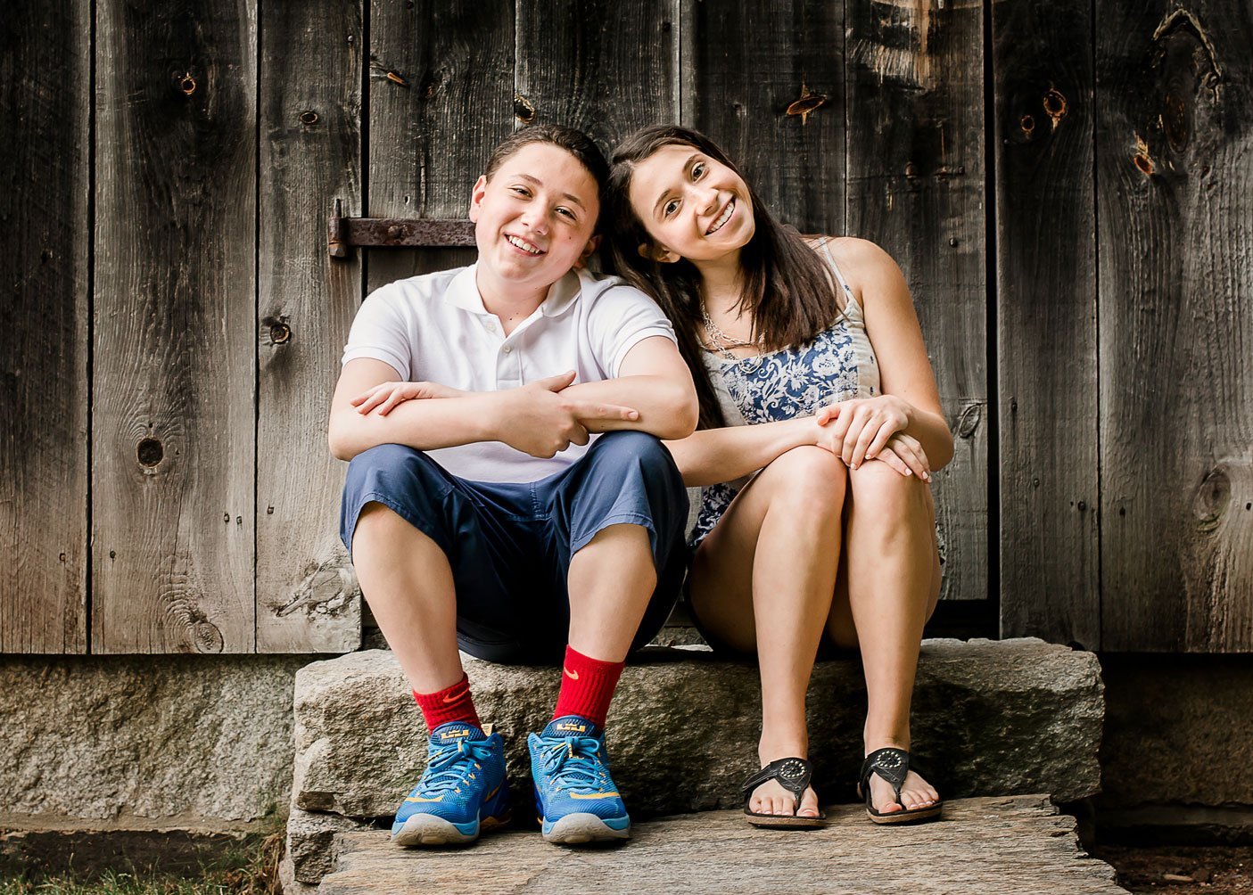 brother and teen sister pose in front of old dark wood barn sitting down with heads together