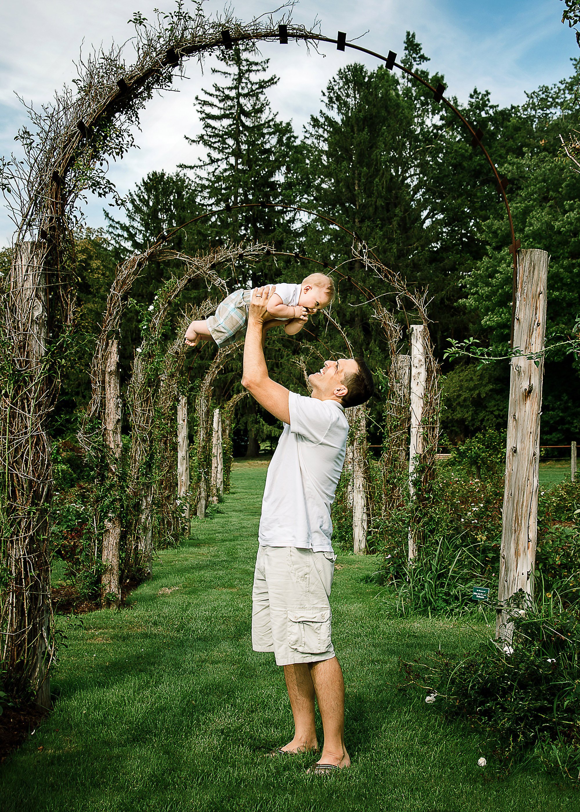 Daddy holds his 6 month old son above his head in a garden underneath arches