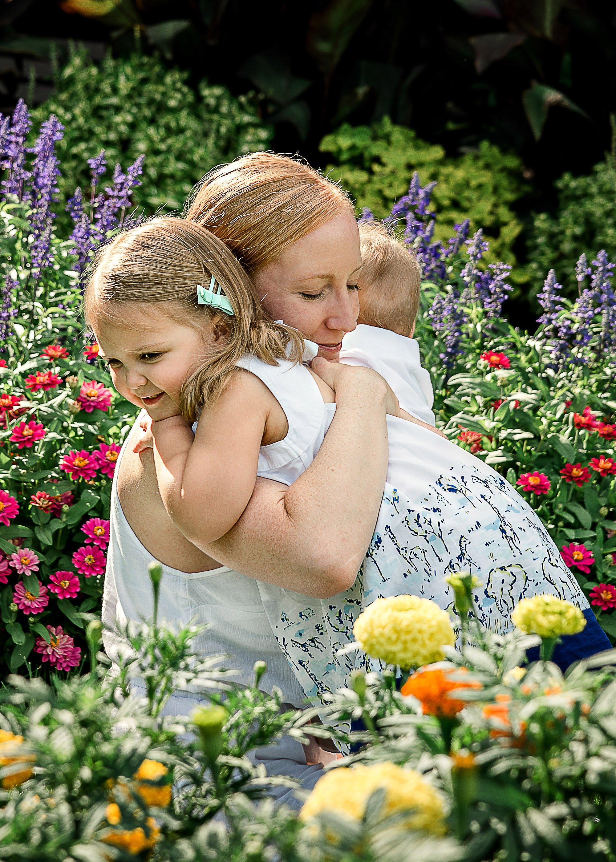 Momma hugging both of her small children in the garden surrounded by flowers