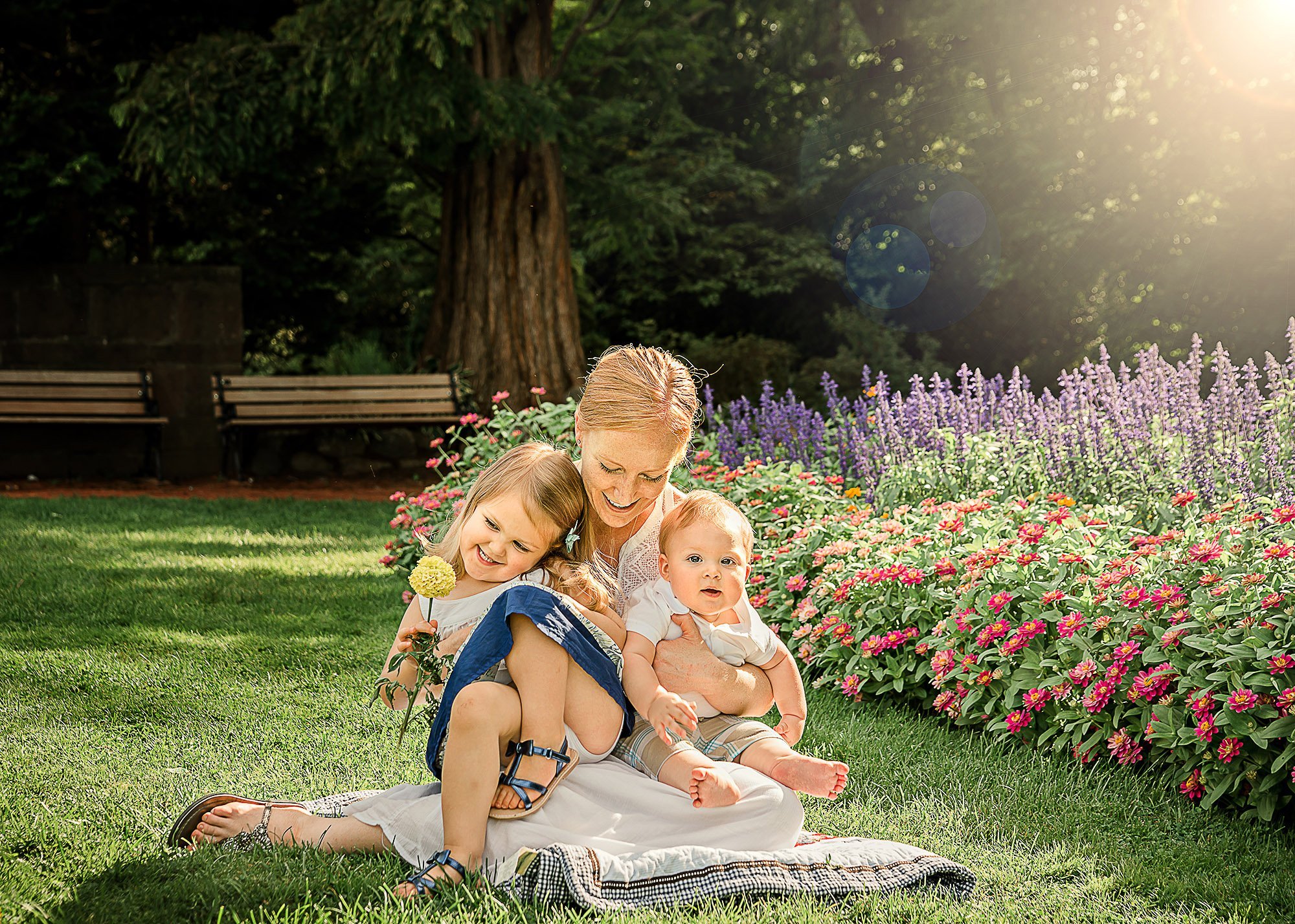 Mother cradles her 2 small children in her lap in the garden with the sunshining down
