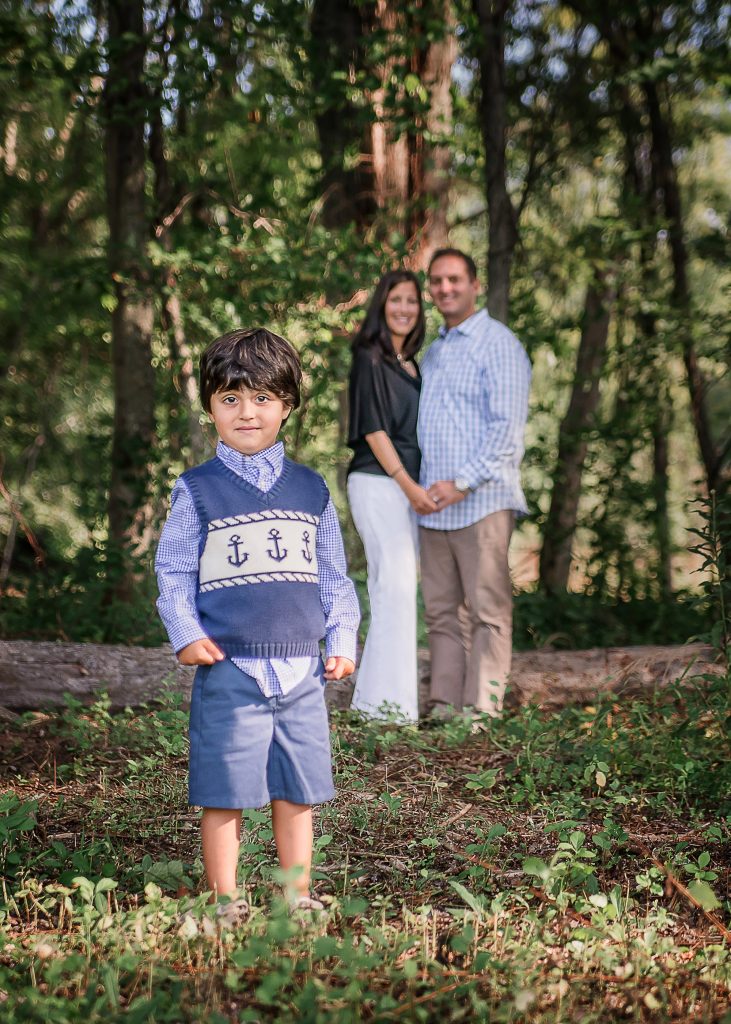 young son stands in front of his parents in the background in the forest in summer