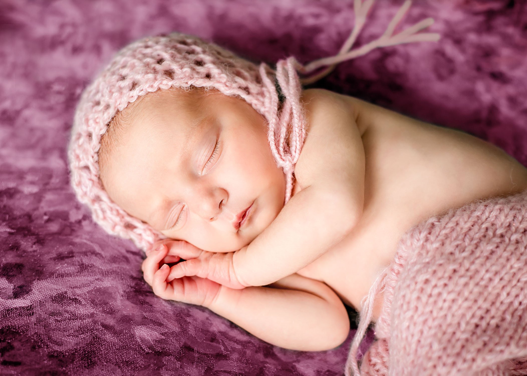 newborn baby girl with pink bonnet and knit pants sleeping on purple bedding