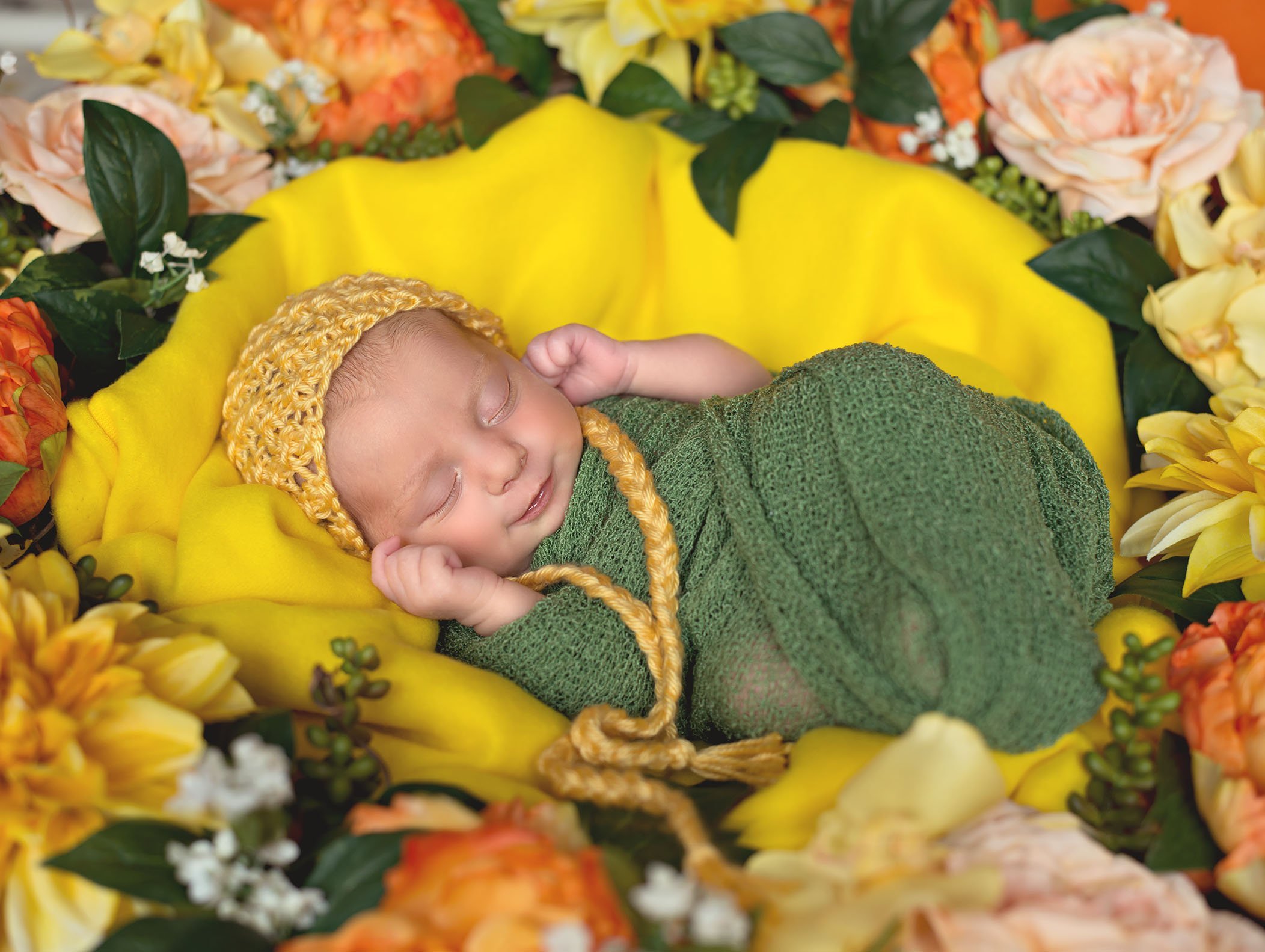 newborn baby girl wrapped in green with a yellow bonnet surrounded by yellow and orange flowers