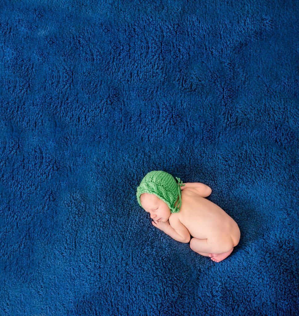 newborn baby in green bonnet sleeping on tummy on big blue blanket
