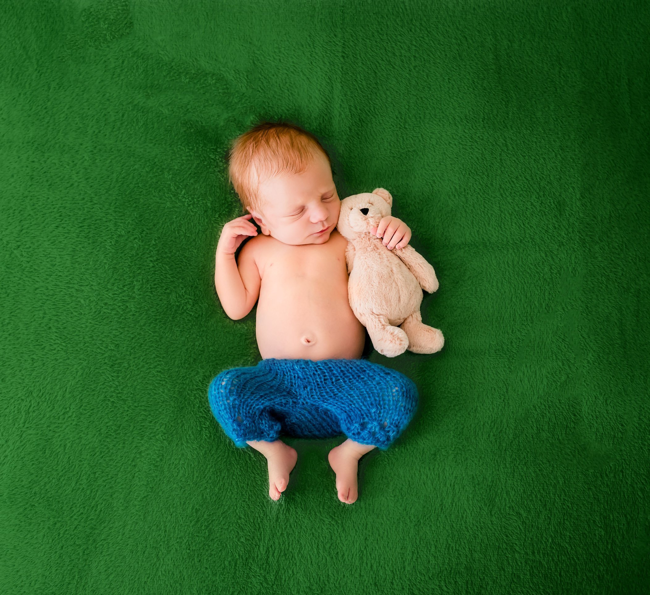 red-headed newborn boy in blue pants sleeping on green blanket with teddy bear