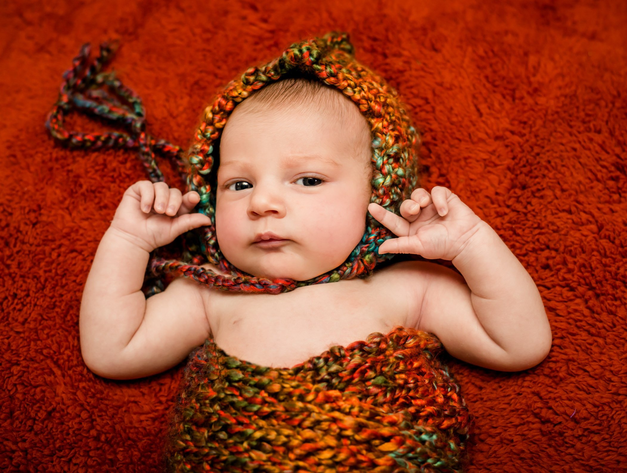 newborn wearing a fall-colored bonnet and wrap on red blanket awake