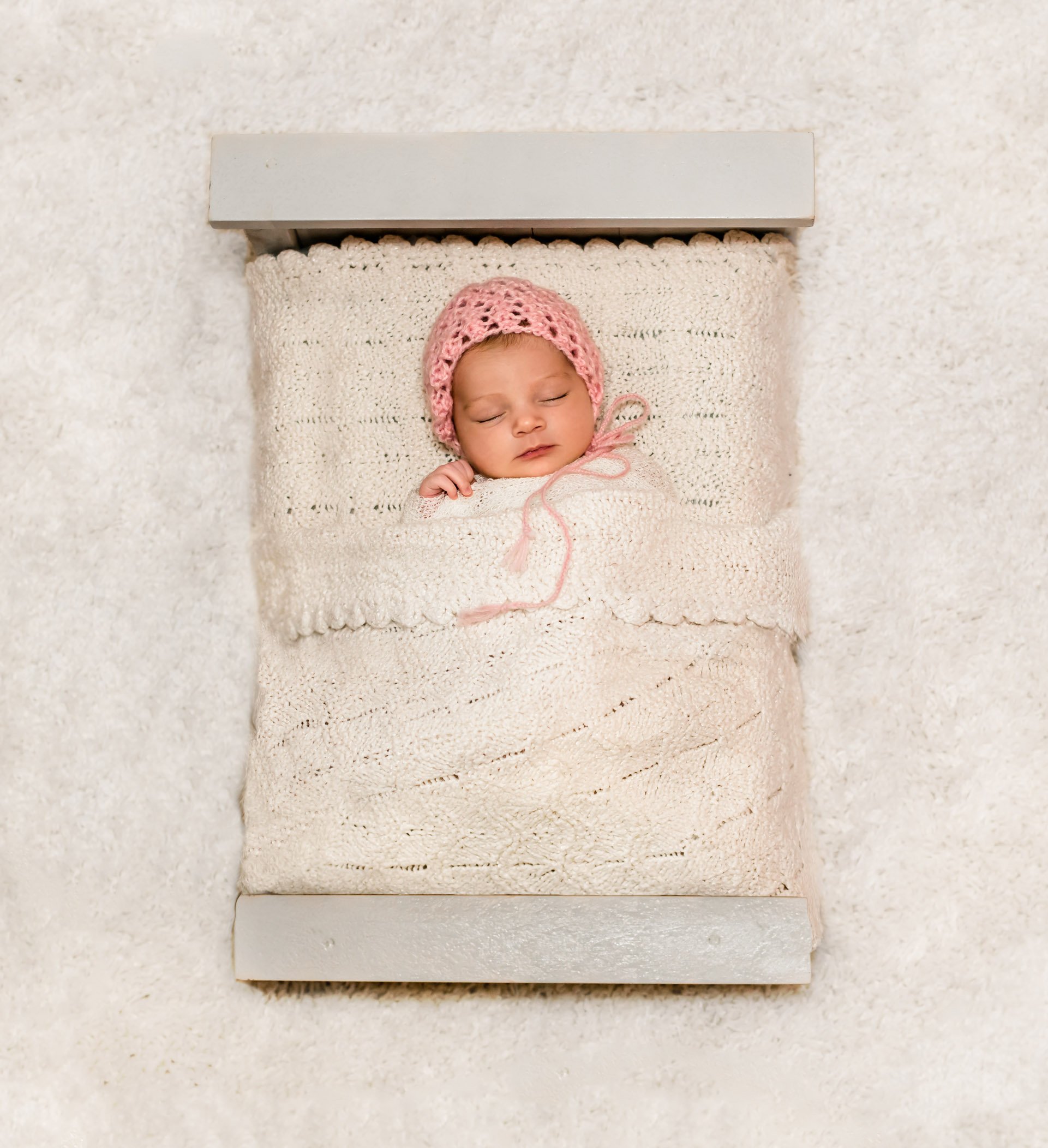 newborn baby girl sleeping in a doll bed with a pink bonnet on
