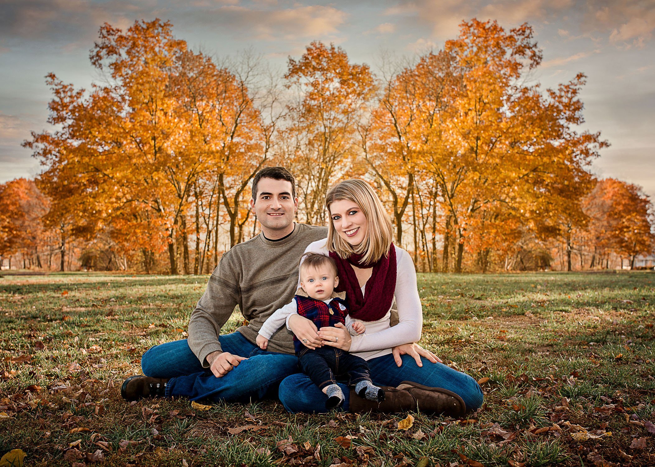 Mom Dad and baby boy sitting outside with orange fall foliage behind them