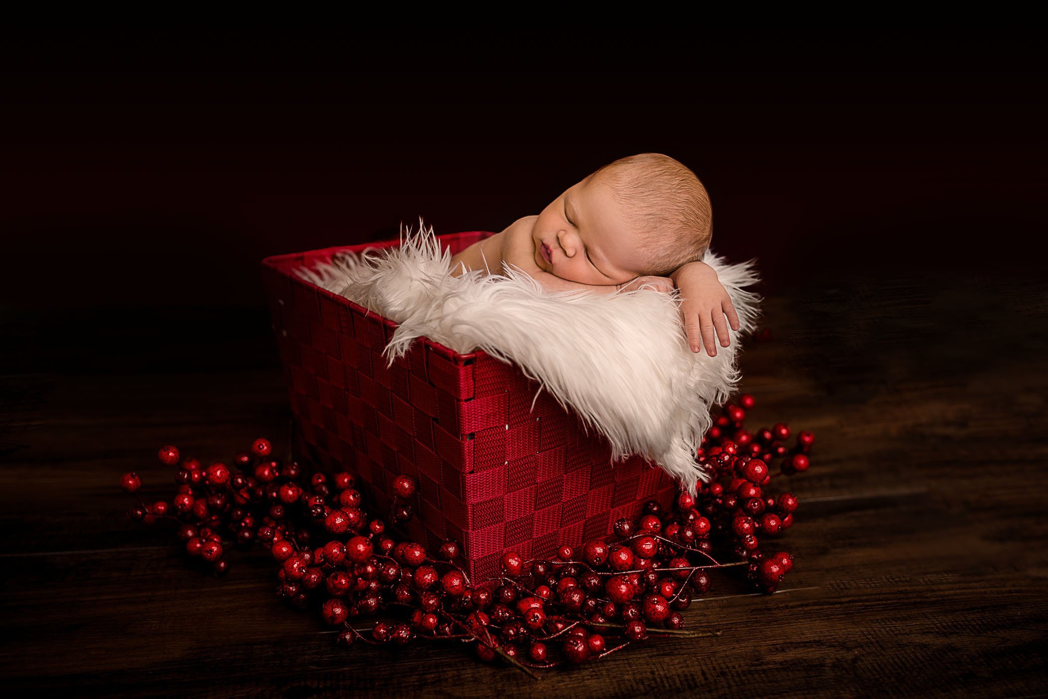 newborn baby sleeping in red basket on white fur with Christmas berries on floor