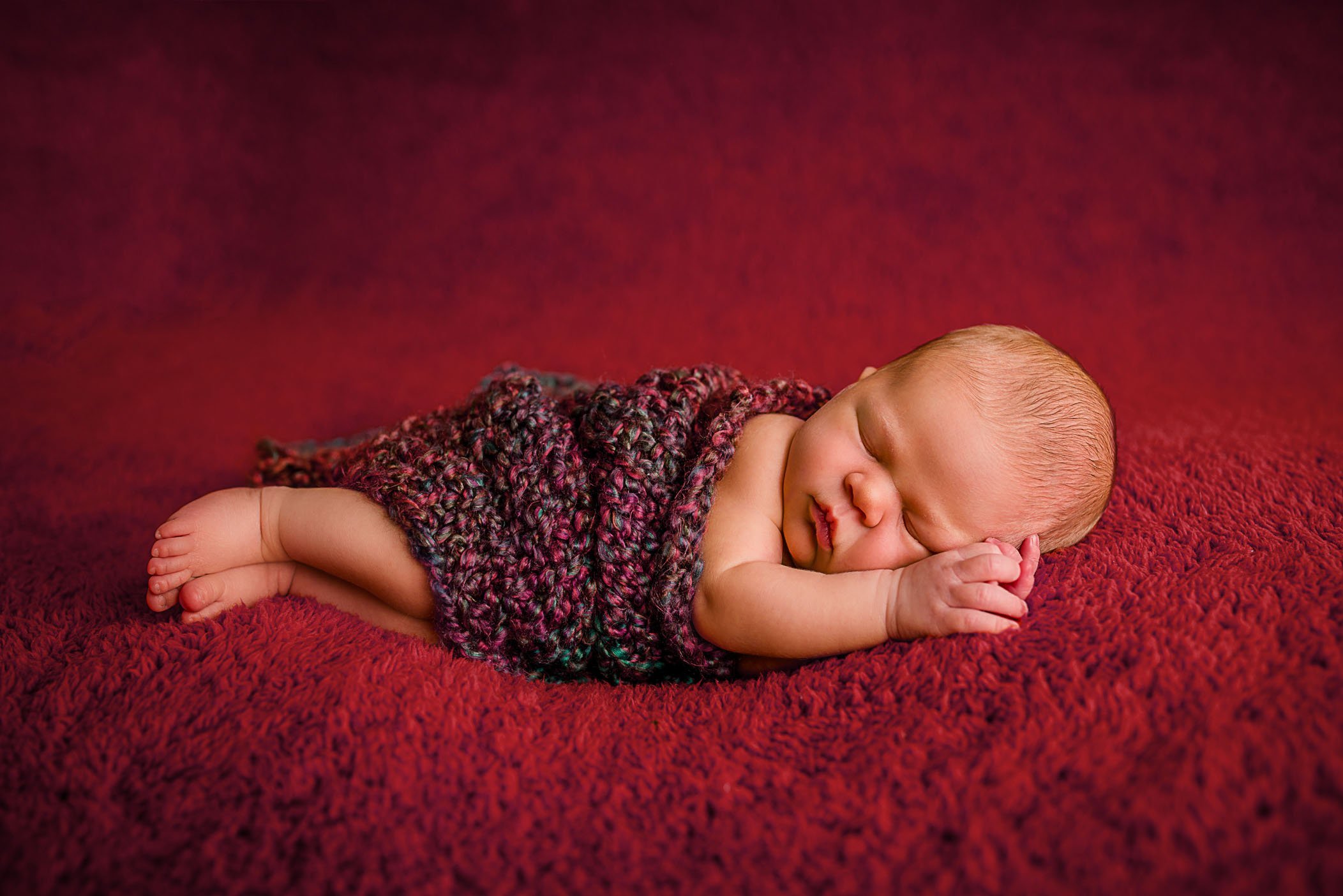 newborn baby sleeping on side on deep red blanket with knitted wrap over her