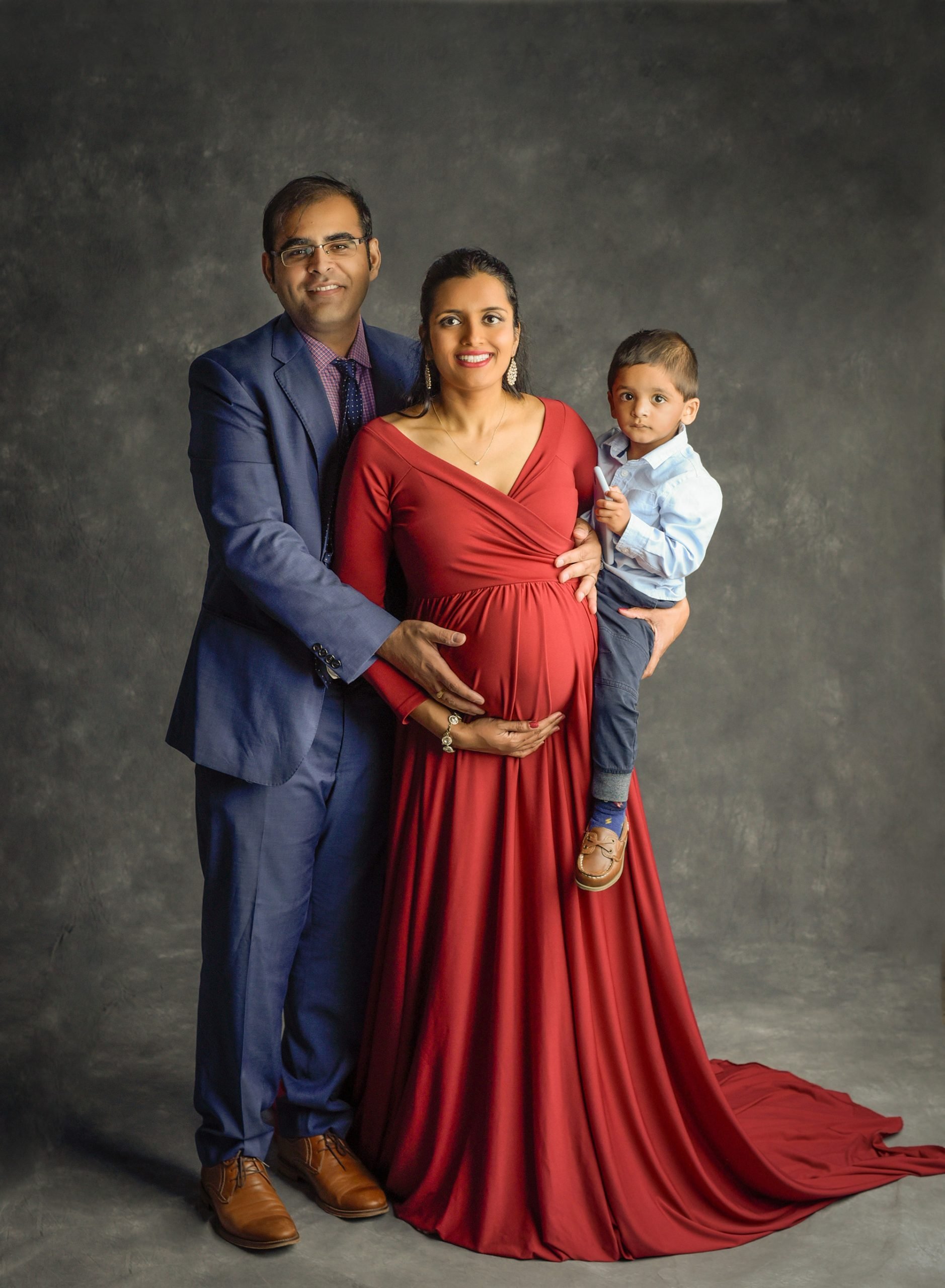 Professional Maternity Newborn Photographer pregnant woman in red maternity dress posing with son and husband